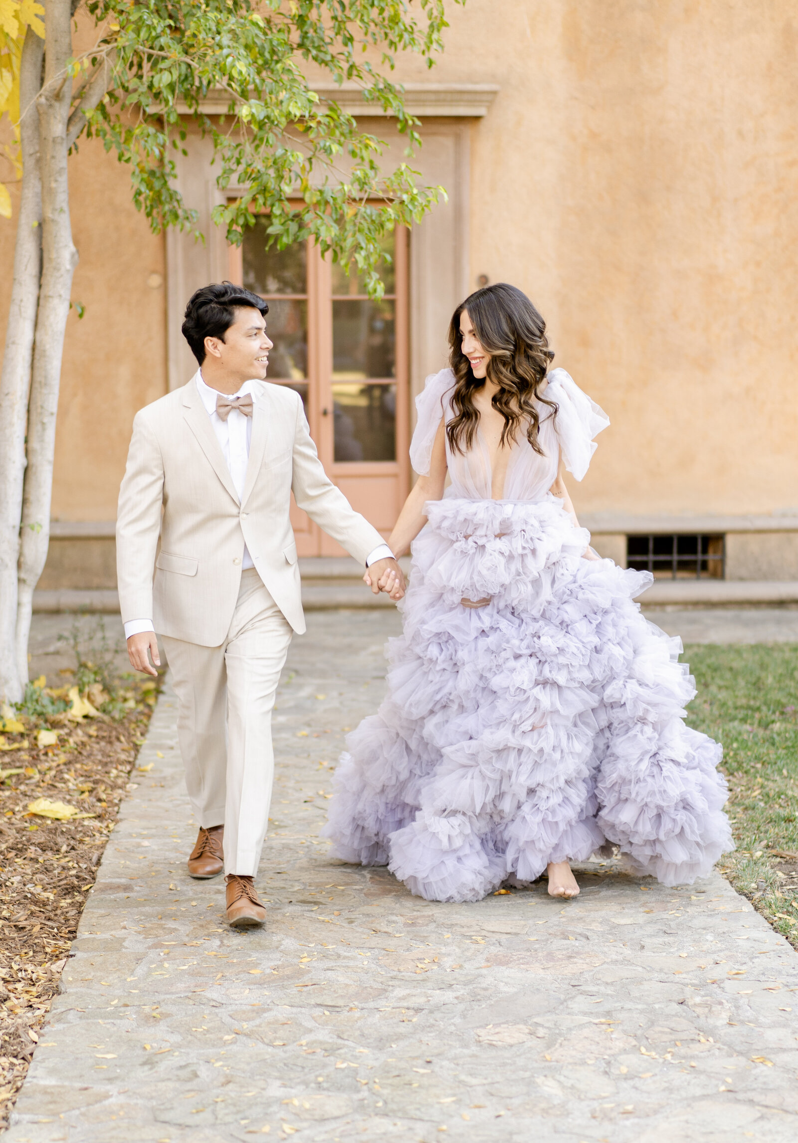 Portrait of bride and groom in a lavender gown and cream suit walking in front of a Spanish-style building with doors.