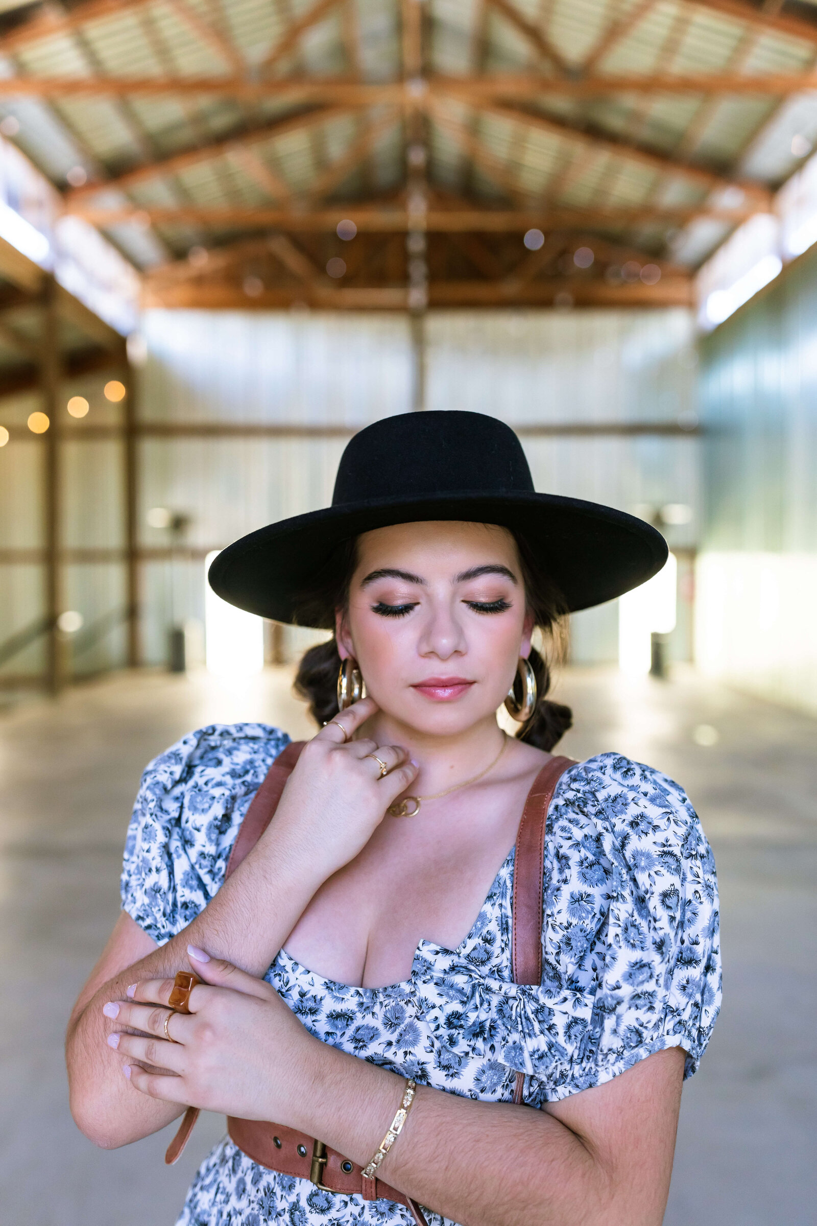 dark haired girl wearing black hat, brown belt, and blue floral dress with bubble braids in a warehouse\