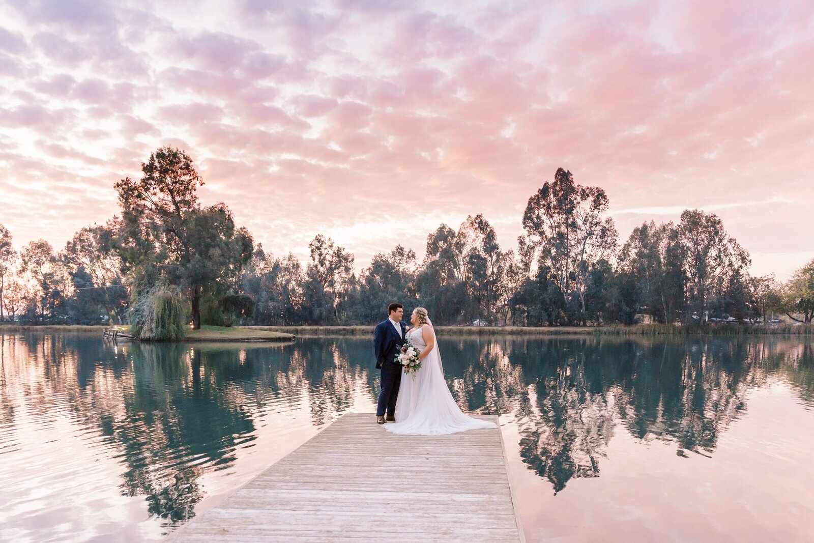 bride and groom standing on a dock by the lake at sunset