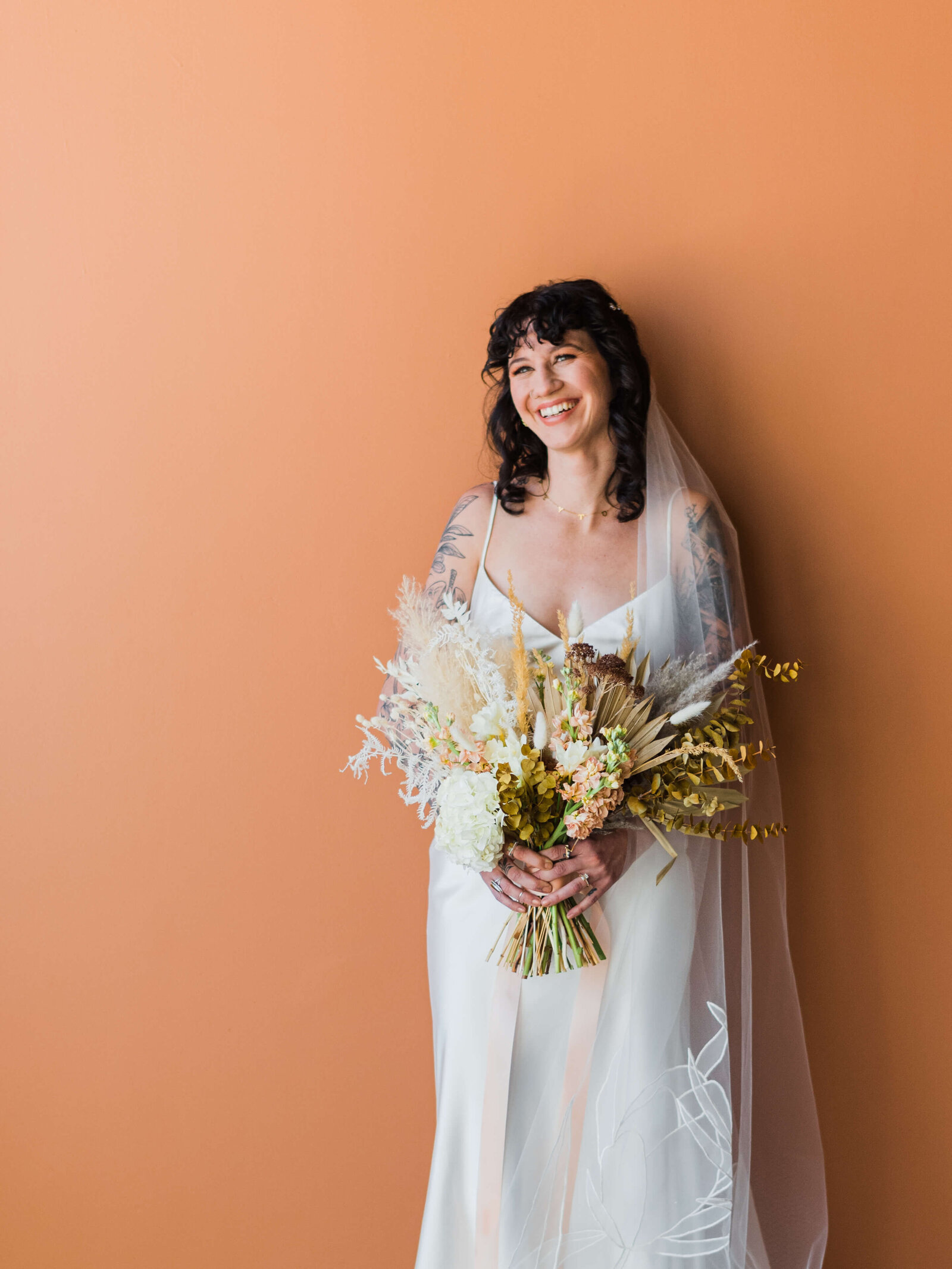 Edgy but beautiful bride in tattooed arms holds a neutrally colored bouquet while standing in front of an orange wall