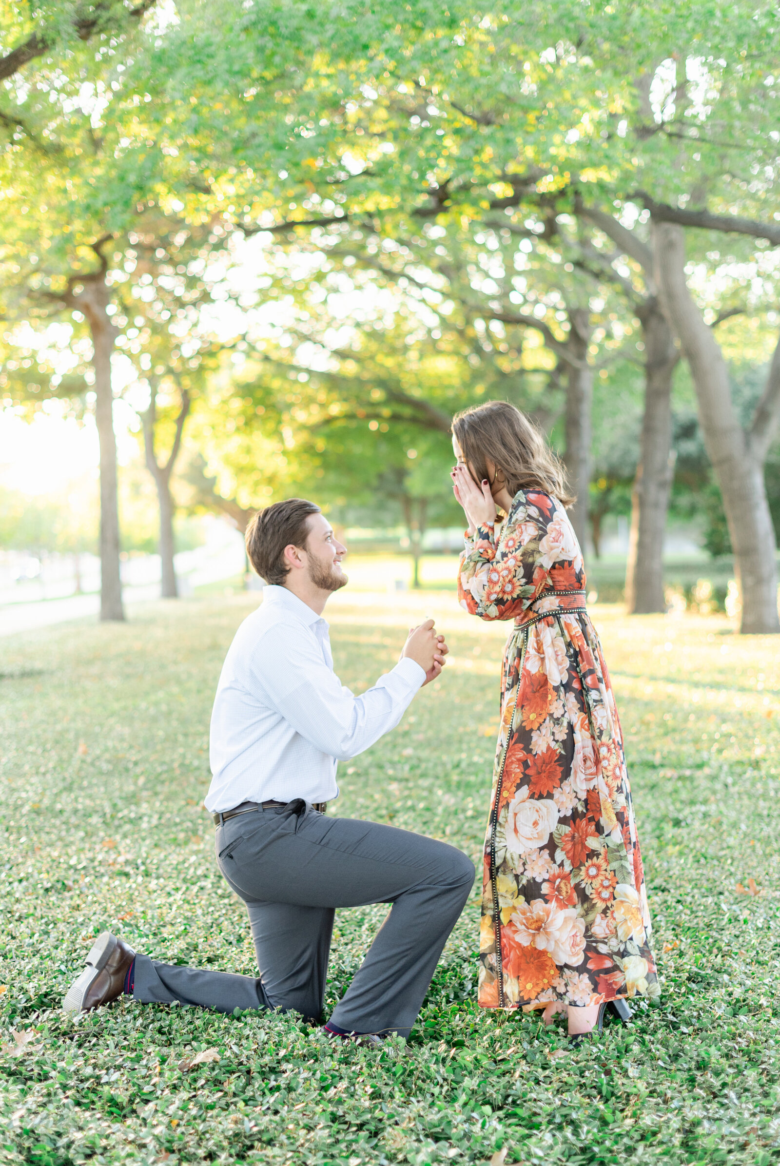 Portrait of a man kneeling and holding a jewewerly box in front of a surprised woman in a floral dress at a park.