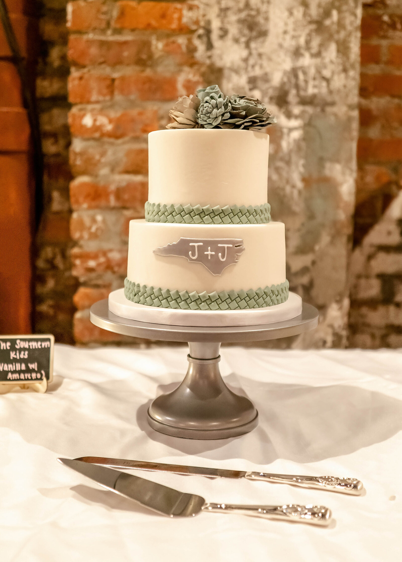 A two-tiered rustic wedding cake with a North Carolina design, sage green braided accents, and pinecone toppers, displayed against a brick backdrop at a Wilmington, NC wedding.