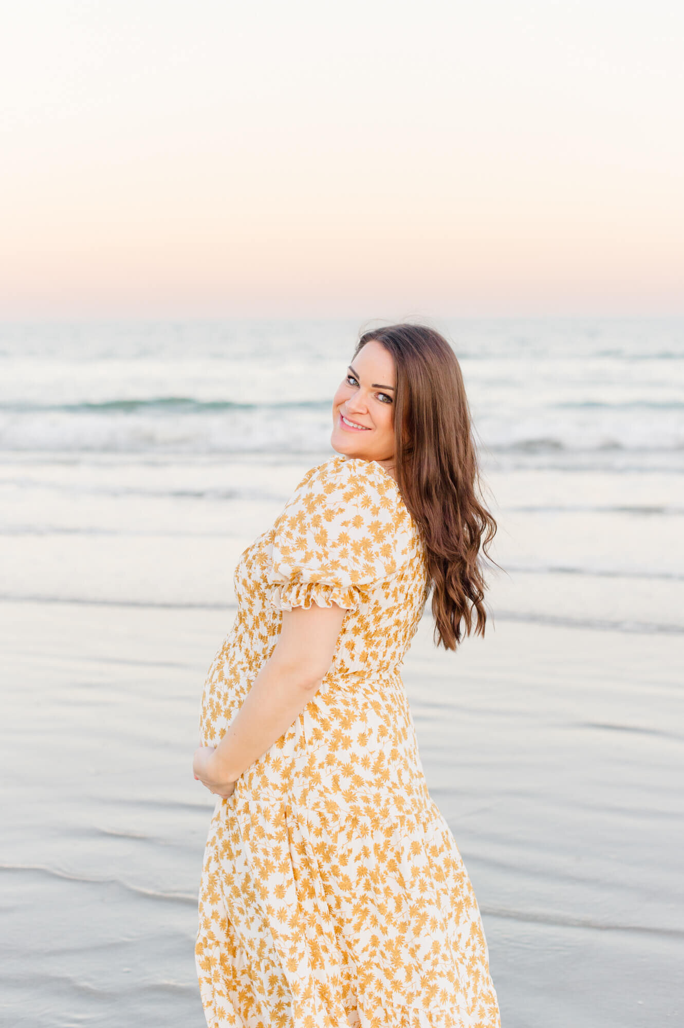 Expectant mother smiles back at the camera while holding her belly on the beach wearing a gorgeous yellow floral dress