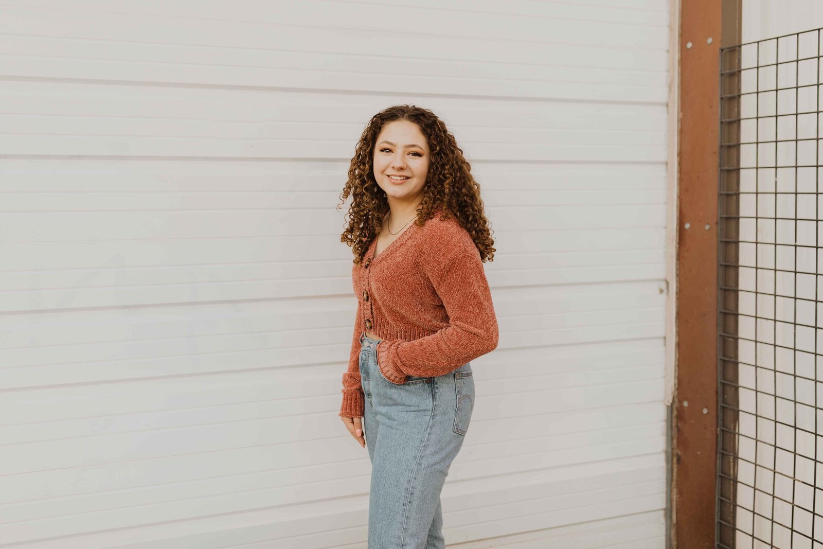 Girl standing in front of white wall, hand in pocket. Wearing a pink sweater and smiling.