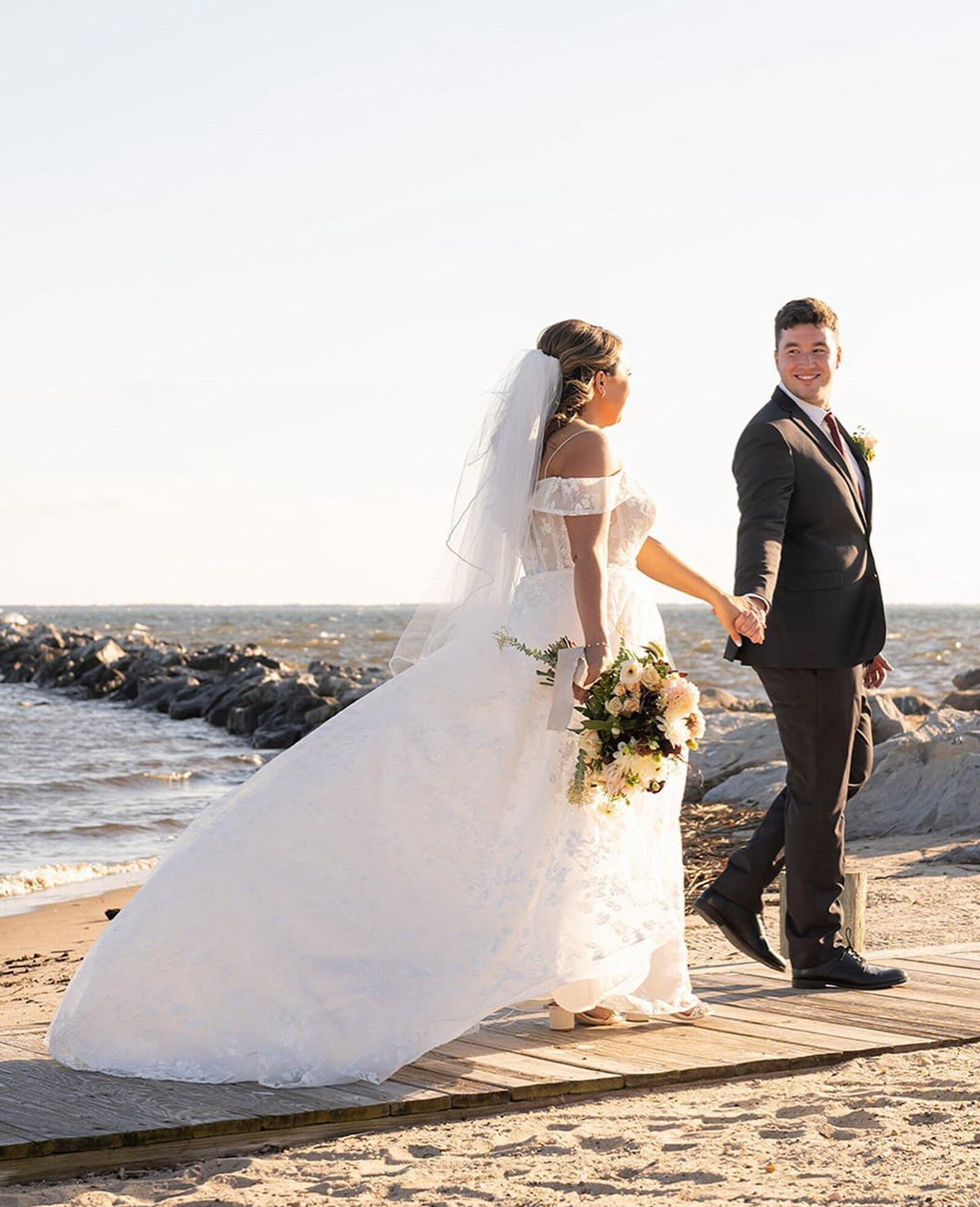 bride weaing a flowing off-shoulder white gown with a long veil and holds a bouquet of flowers and groom in a dark suit and tie walking hand-in-hand along a boardwalk by the sea
