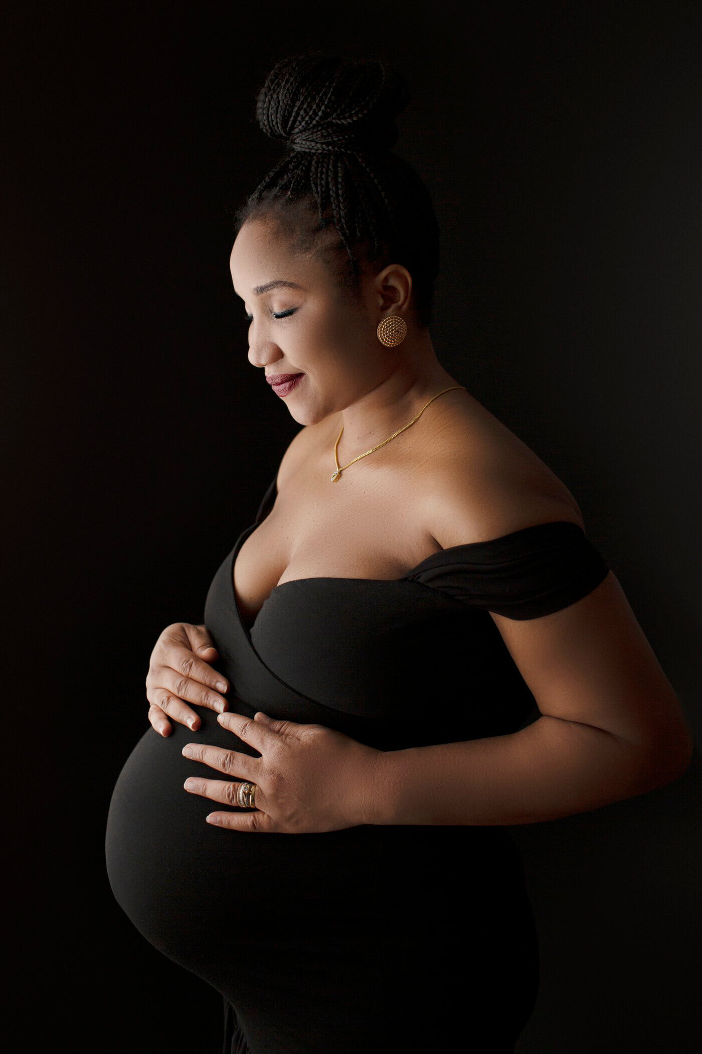 A pregnant woman stands against a black background, cradling her baby bump with both hands. She is wearing an elegant, off-the-shoulder black dress that beautifully accentuates her figure. Her hair is styled in a high braided bun, and she is adorned with gold earrings and a delicate gold necklace. The lighting softly illuminates her profile, highlighting her serene and content expression. The image captures a moment of quiet anticipation and grace, with the dark background creating a striking contrast against her radiant skin and elegant attire.