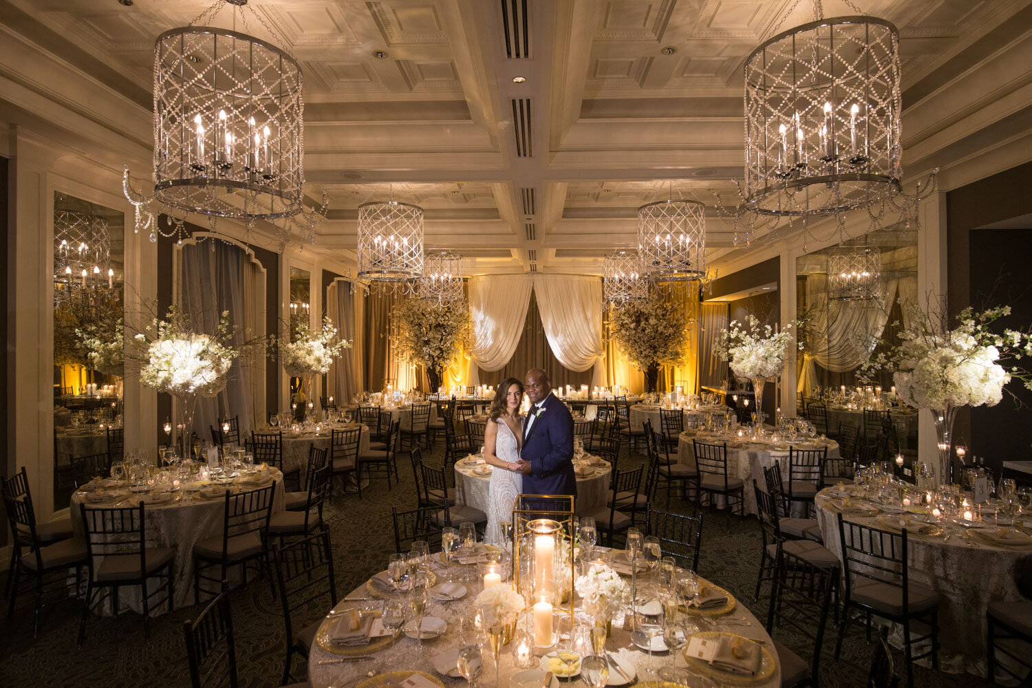 A bride and groom are standing in a large room with tables set for a wedding reception.