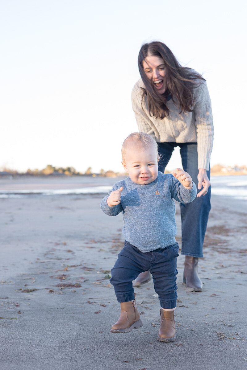 Smiling toddler running on the beach with mom laughing behind him during a playful family photo session in Connecticut. Captured by Nicole, a lifestyle family photographer for joyful, relaxed families.