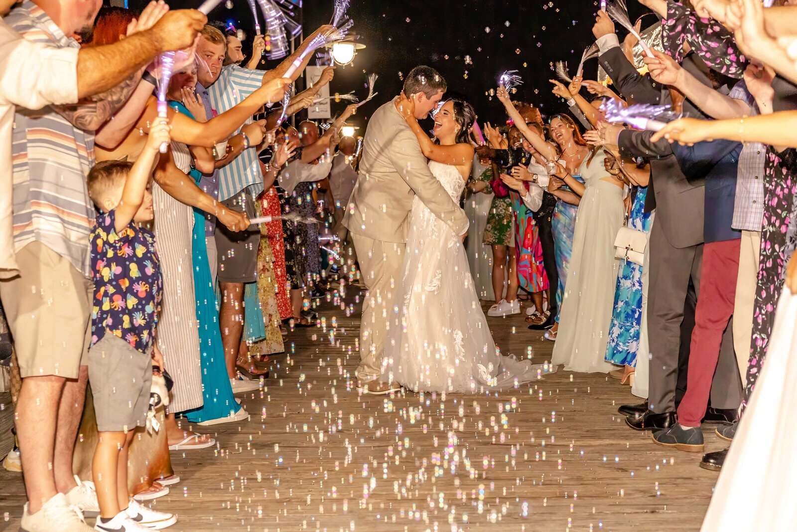The bride and groom share a kiss during a joyful sparkler exit surrounded by guests, captured at a Topsail Beach, NC wedding, showcasing a fun and celebratory moment.