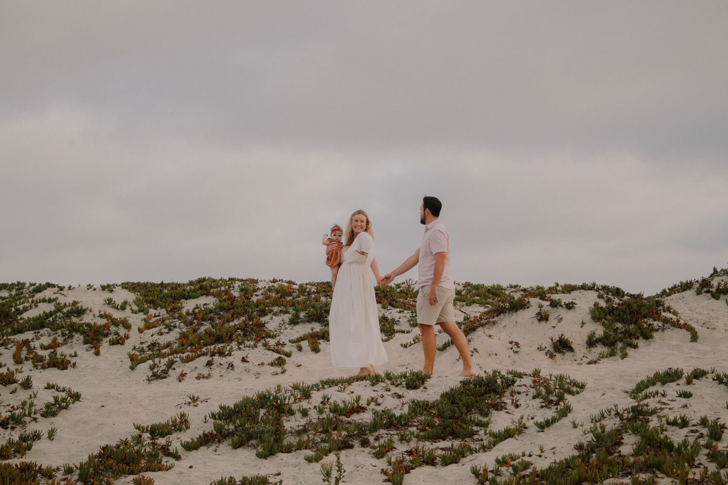 Family of three walking on a sandy hill, with parents holding their baby, captured by a San Diego family photographer