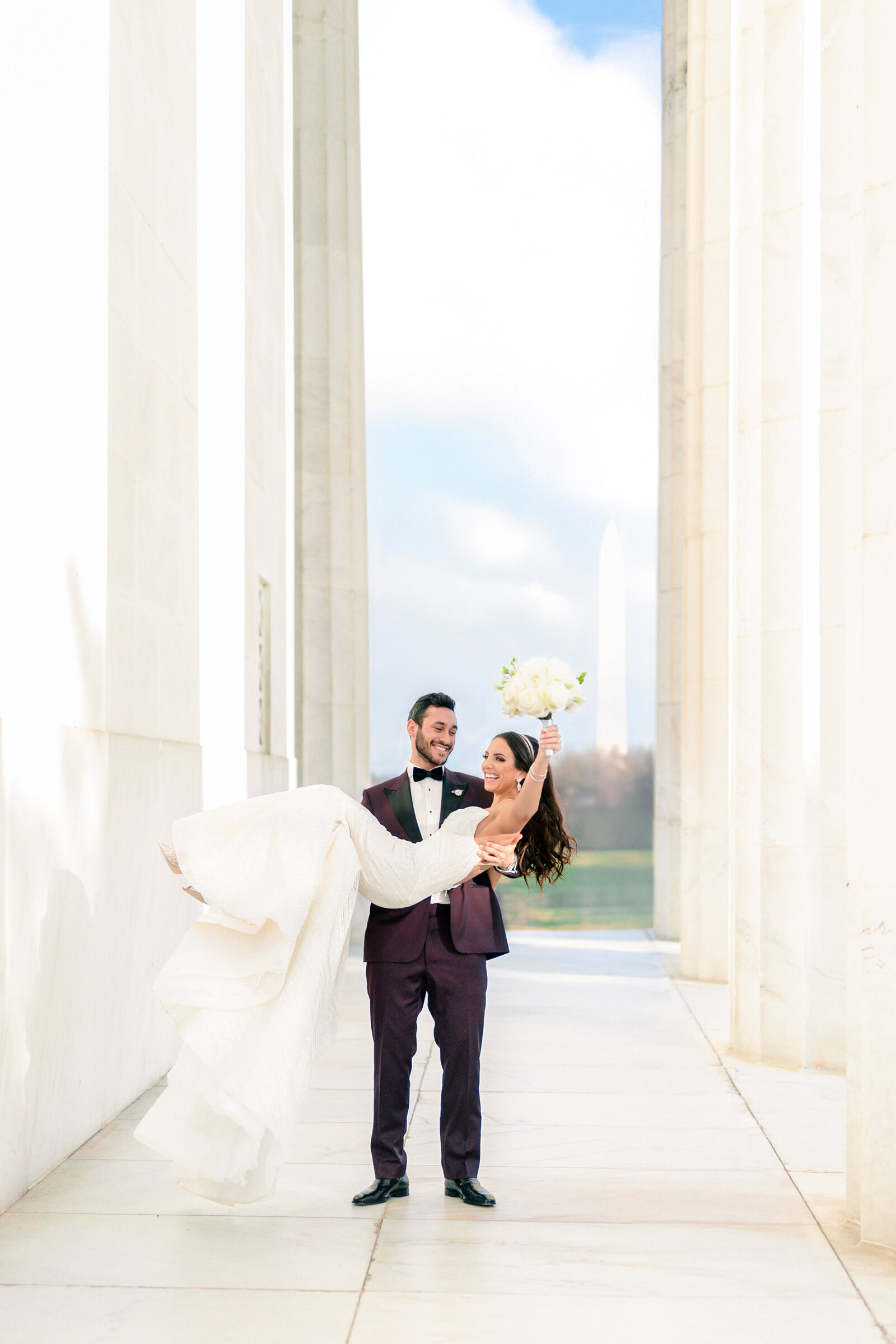 A groom in a tuxedo joyfully carries his bride in a white dress along a sunlit corridor. The bride holds a bouquet and smiles, with a backdrop of tall columns and a distant view of a monument.