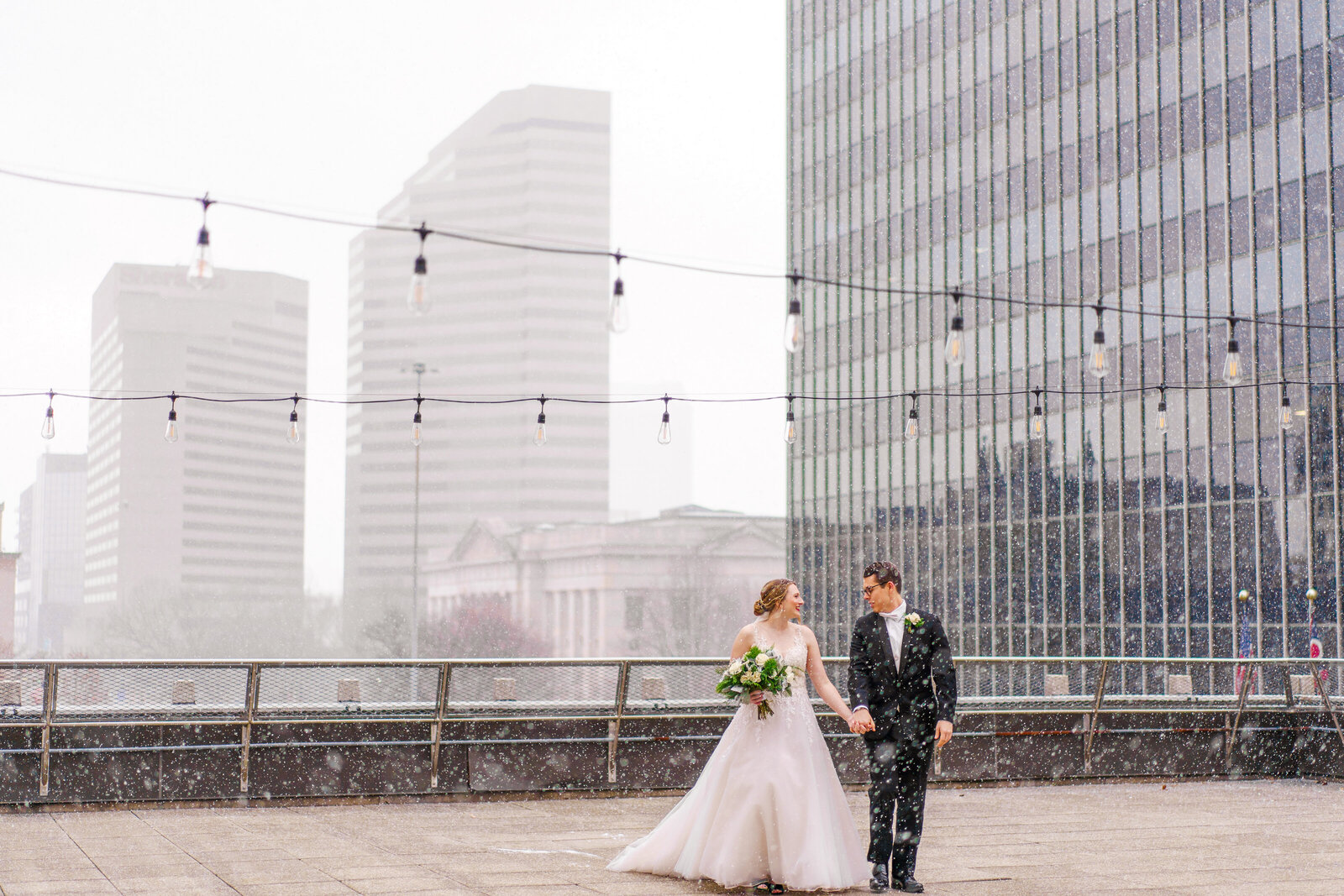 Bride and groom hold hands, look at each other, and walk on a rooftop in downtown Columbus as they're showered with snow. December wedding.