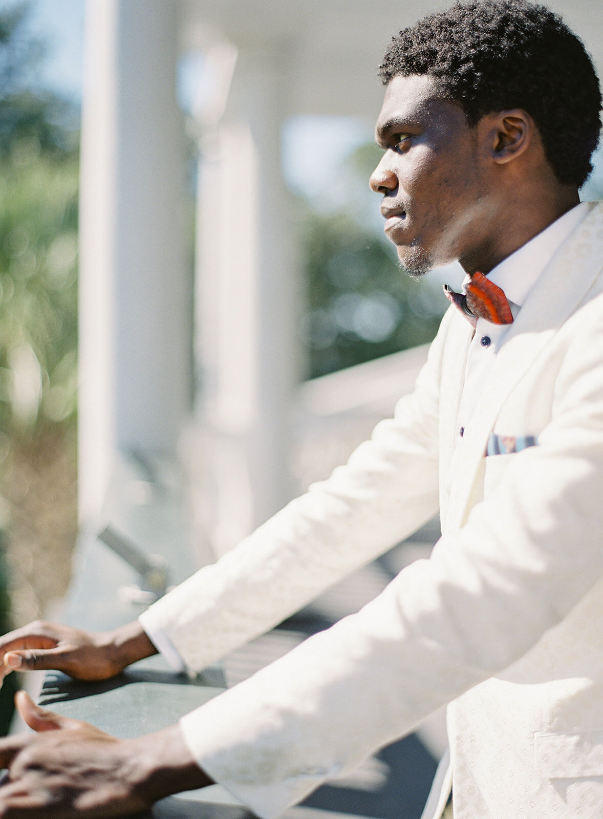 Profile of groom from his hips up. He is wearing an ivory and gold tuxedo with his arms stretched out and resting on a balcony railing and he is looking out the the front yard. Photographed at Lowndes Grove by wedding photographers in Charleston Amy Mulder Photography.