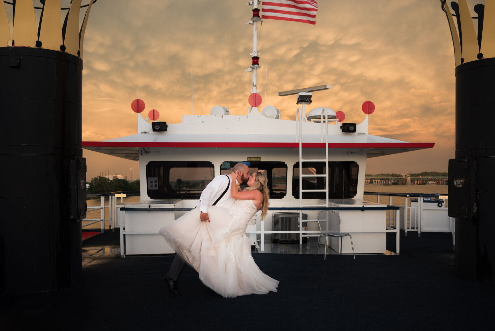 Groom dips bride on river boat under sunset and flag