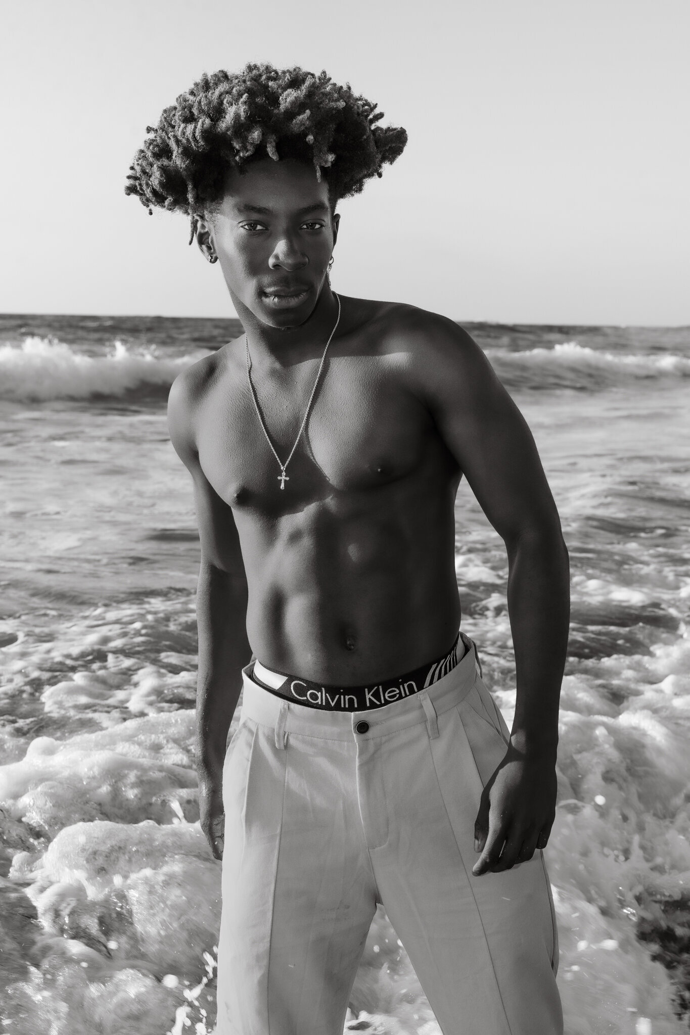 Black and white snapshot of male standing in the water at a beach wearing white trousers and calvin klein boxer label showing
