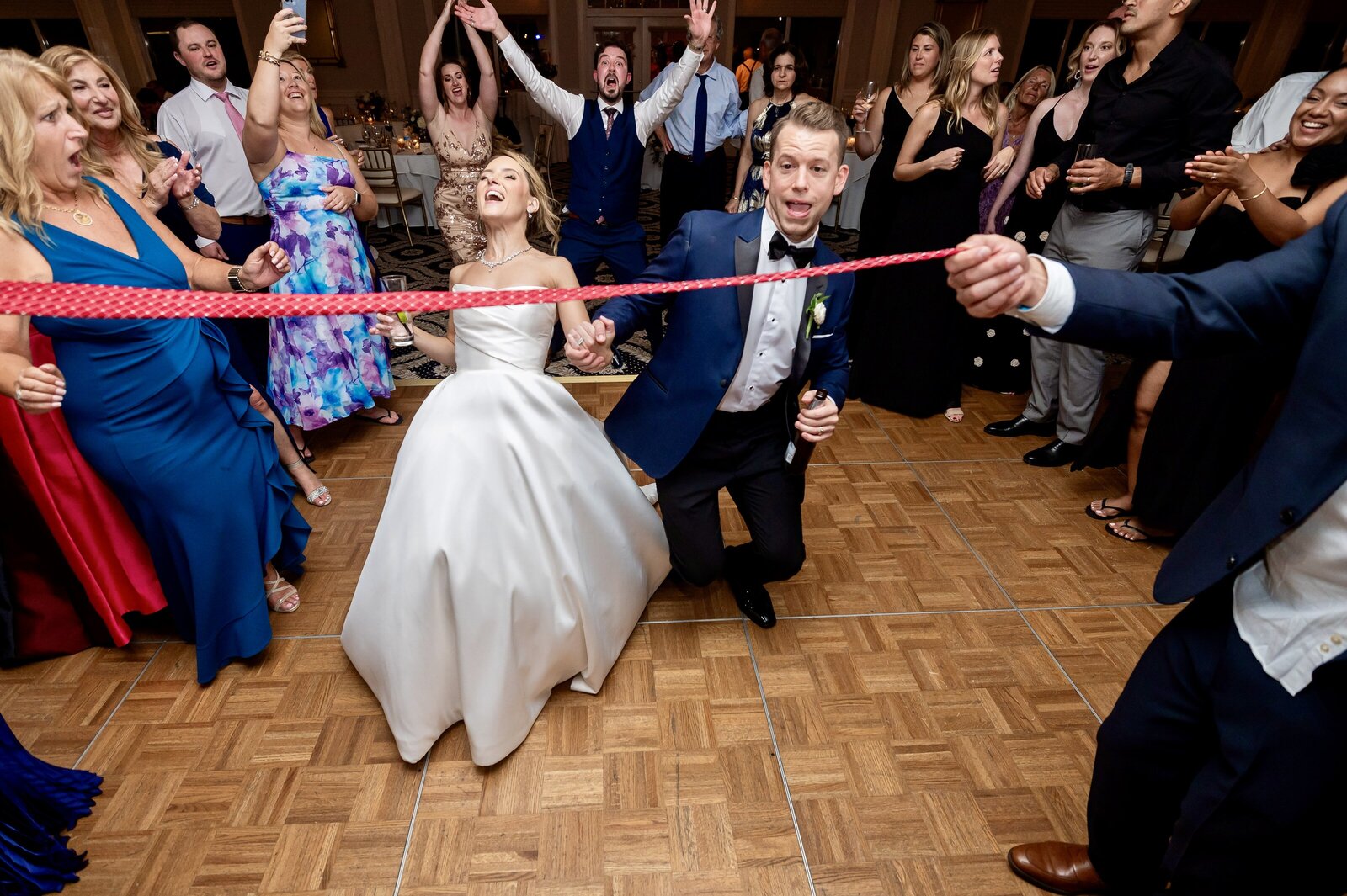 A bride and groom limbo under a red ribbon on a dance floor, surrounded by cheering guests.