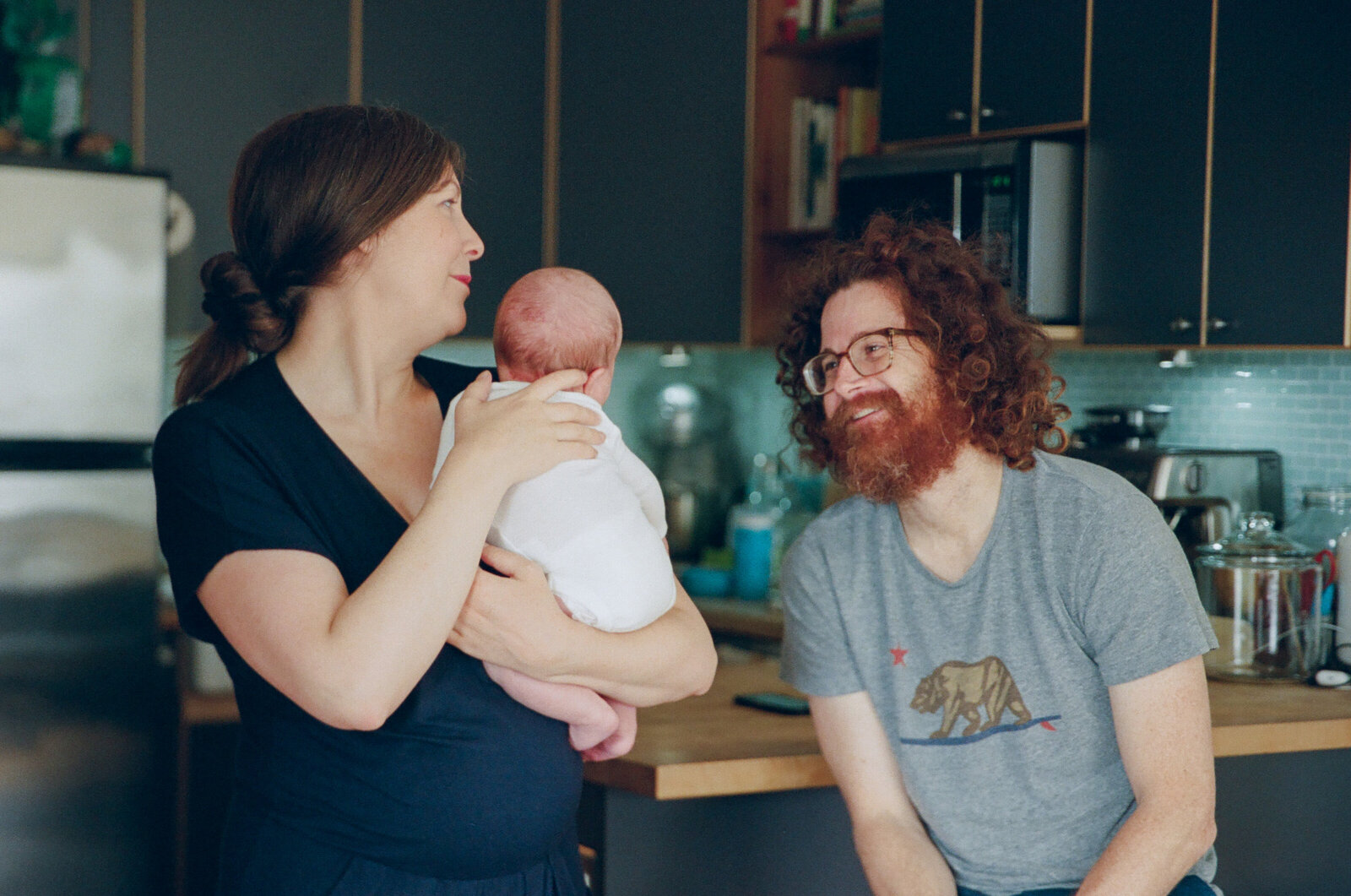 mom in navy jumpsuit holding newborn baby girl while dad with curly red hair and beard smiles happily at them