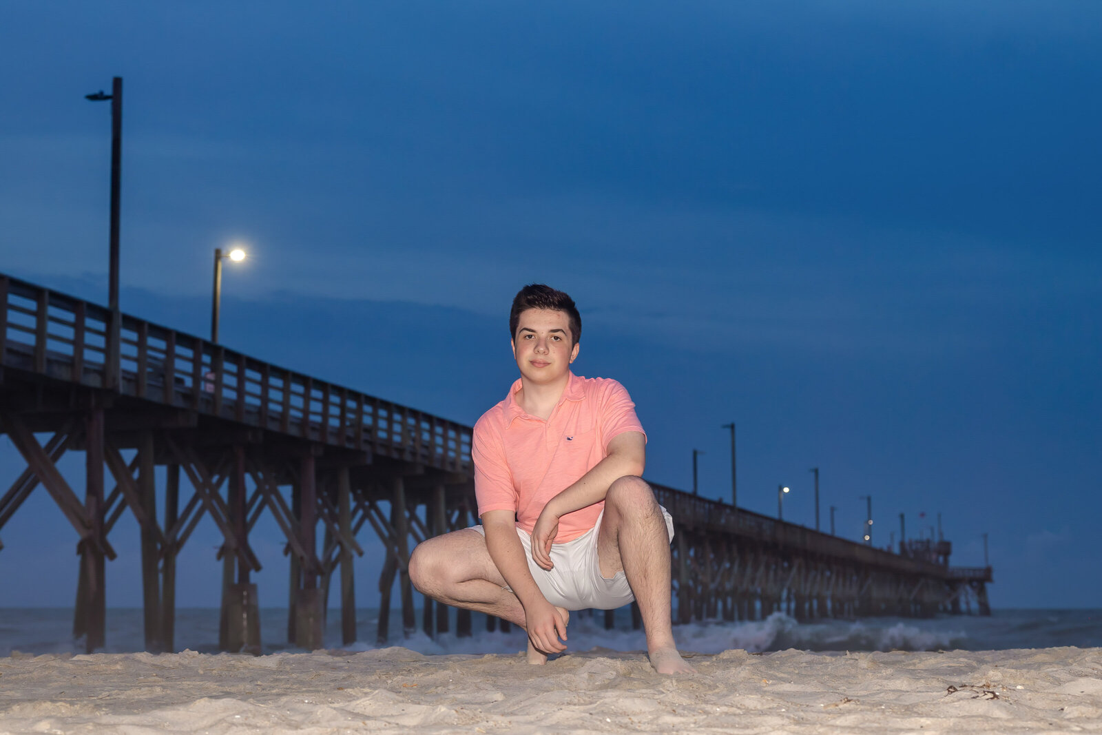 A high school senior in a pink shirt and shorts crouching on the beach at night with a pier illuminated behind in North Carolina. This atmospheric portrait captures the unique charm of senior photography, perfect for evening beach sessions.