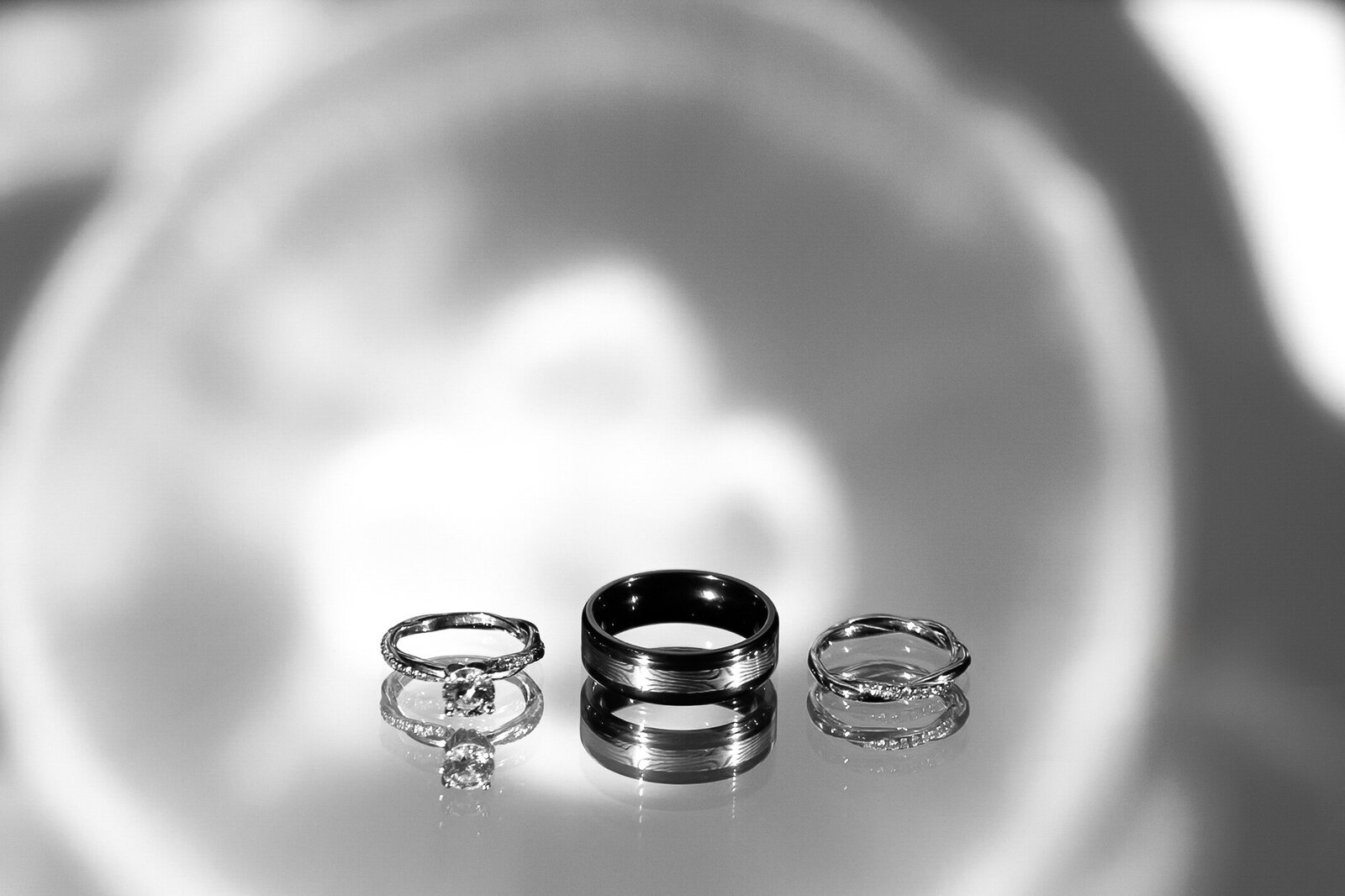 A black-and-white photo of three wedding rings displayed on a reflective surface with a blurred background, captured during a Pine Knoll Shores Aquarium wedding in NC, showcasing timeless and elegant wedding details.