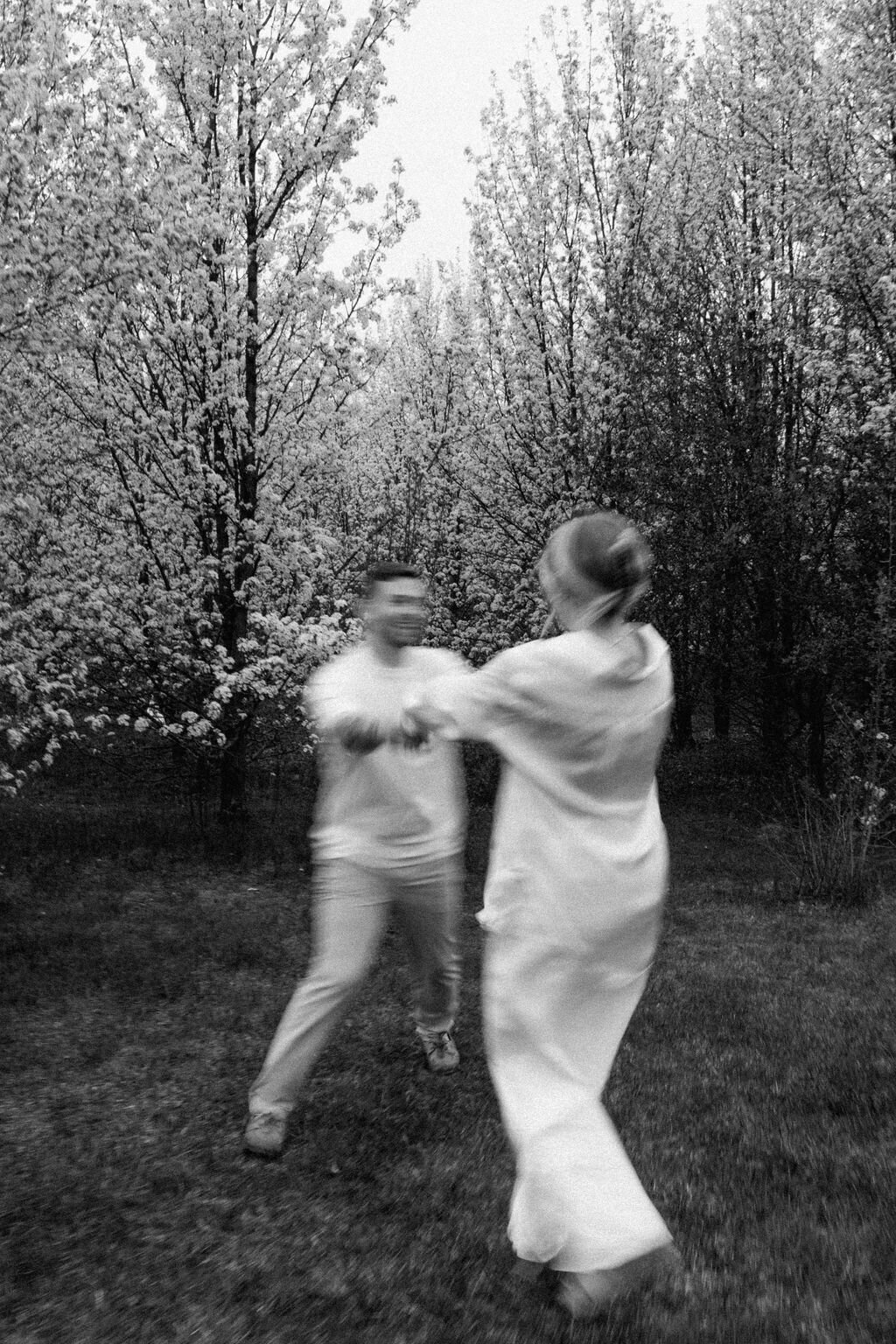 Black and white photo of couple in white holding both hands and spinning together surrounded by cherry blossom trees