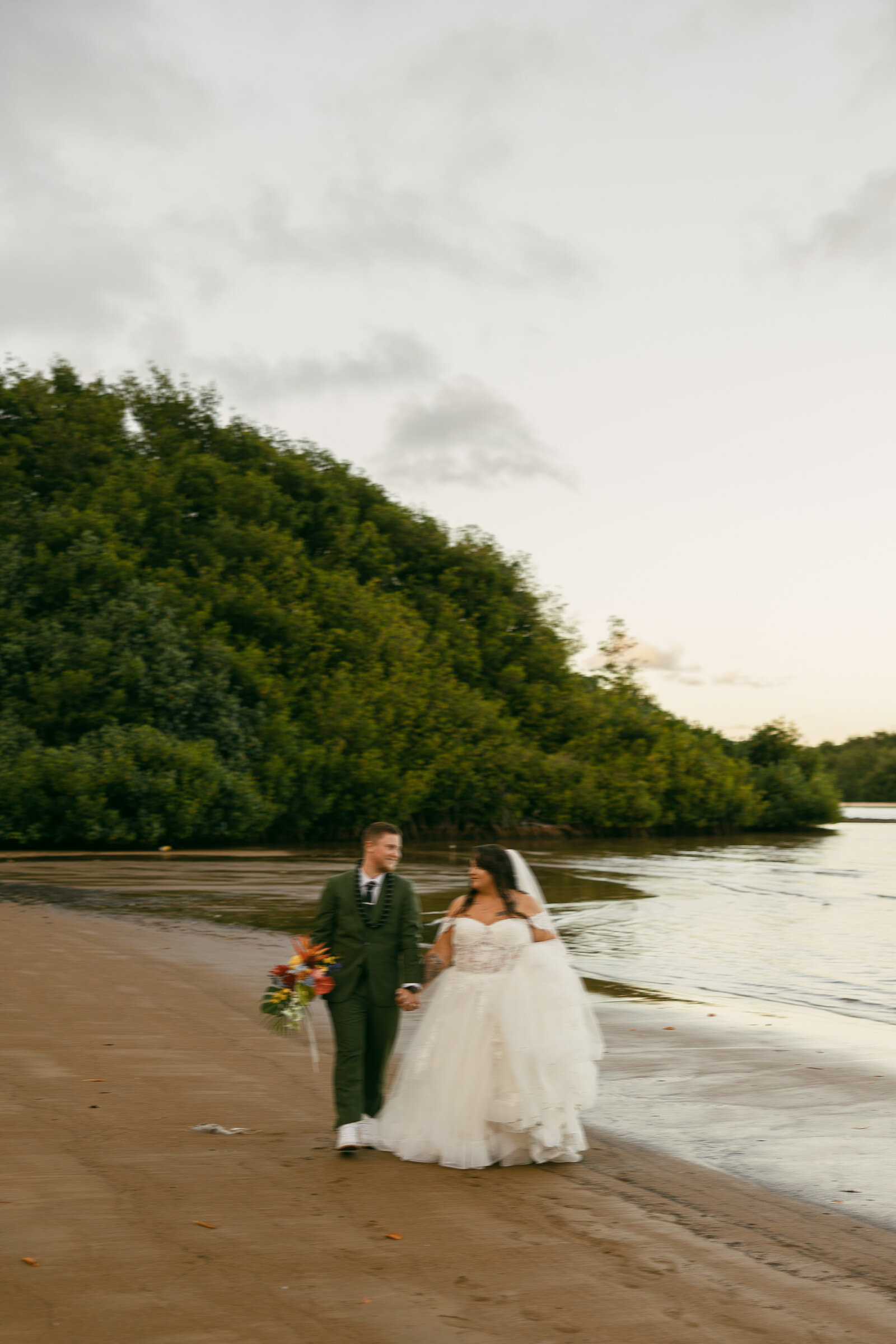 A bride and groom holding hands and walking along the water's edge.