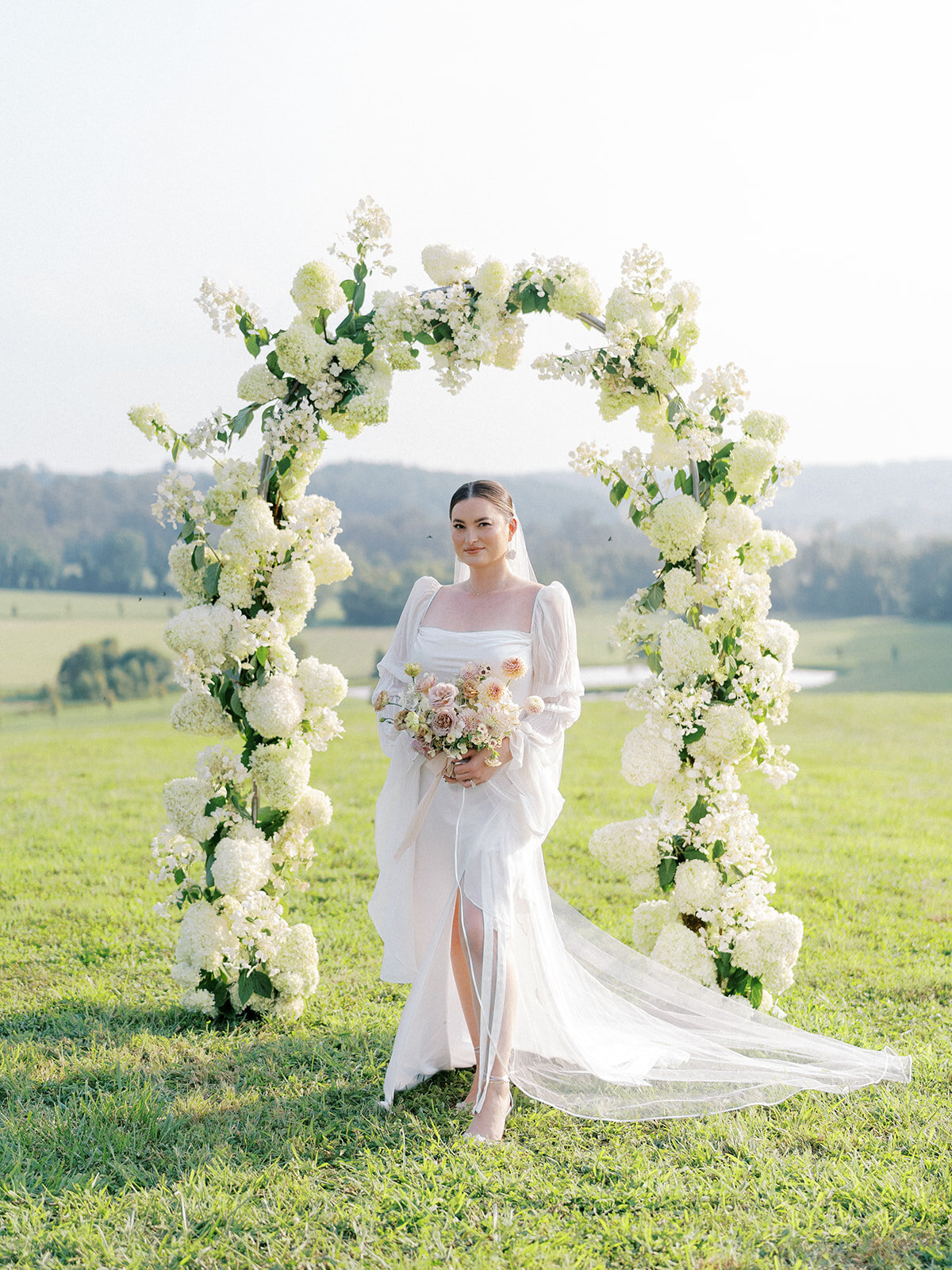 Portrait of bride in front of a full coverage floral arch filled with white airy quick-fire hydrangea and limelight hydrangeas