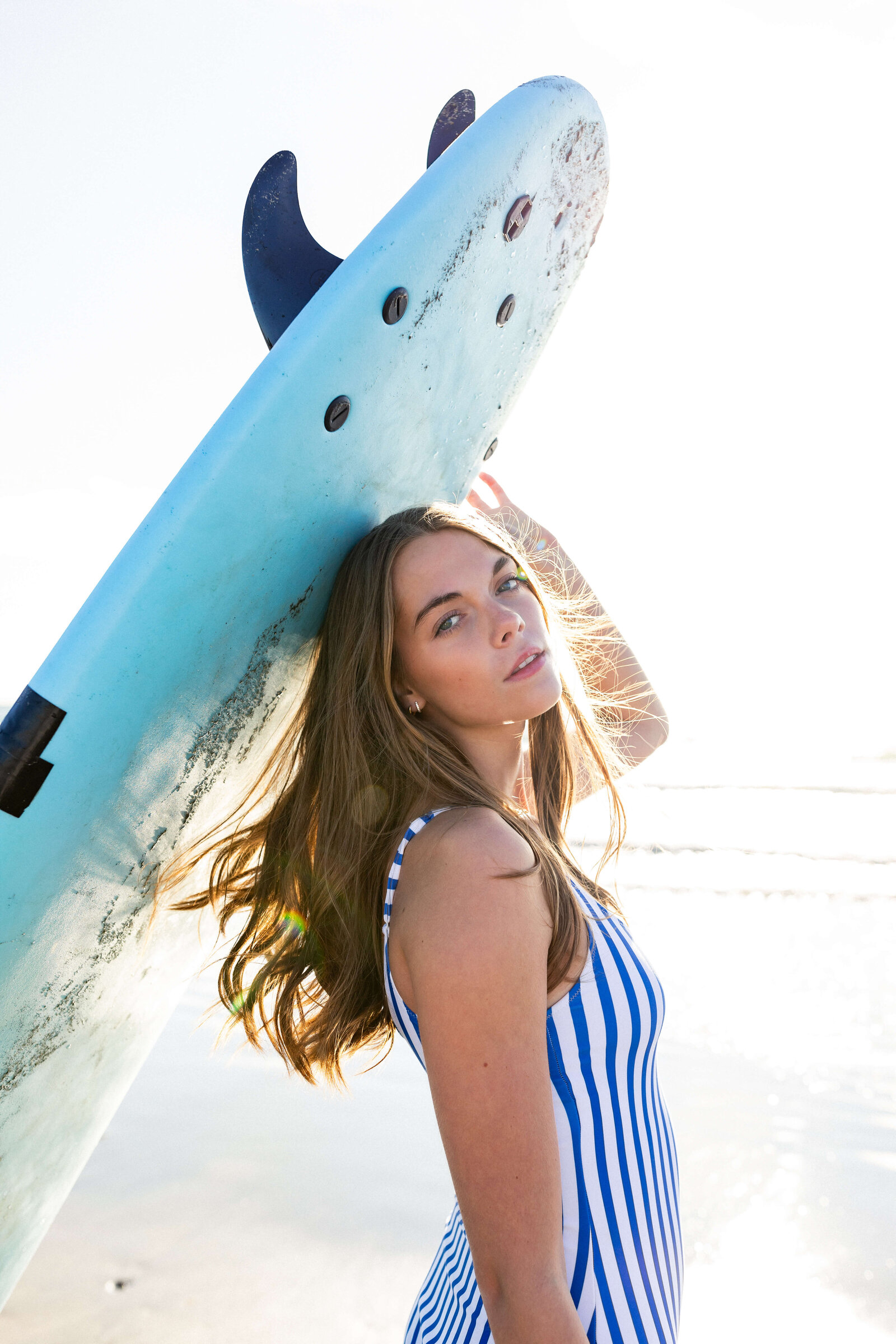 brunette girl holding a surf board wearing a striped one piece swimsuit