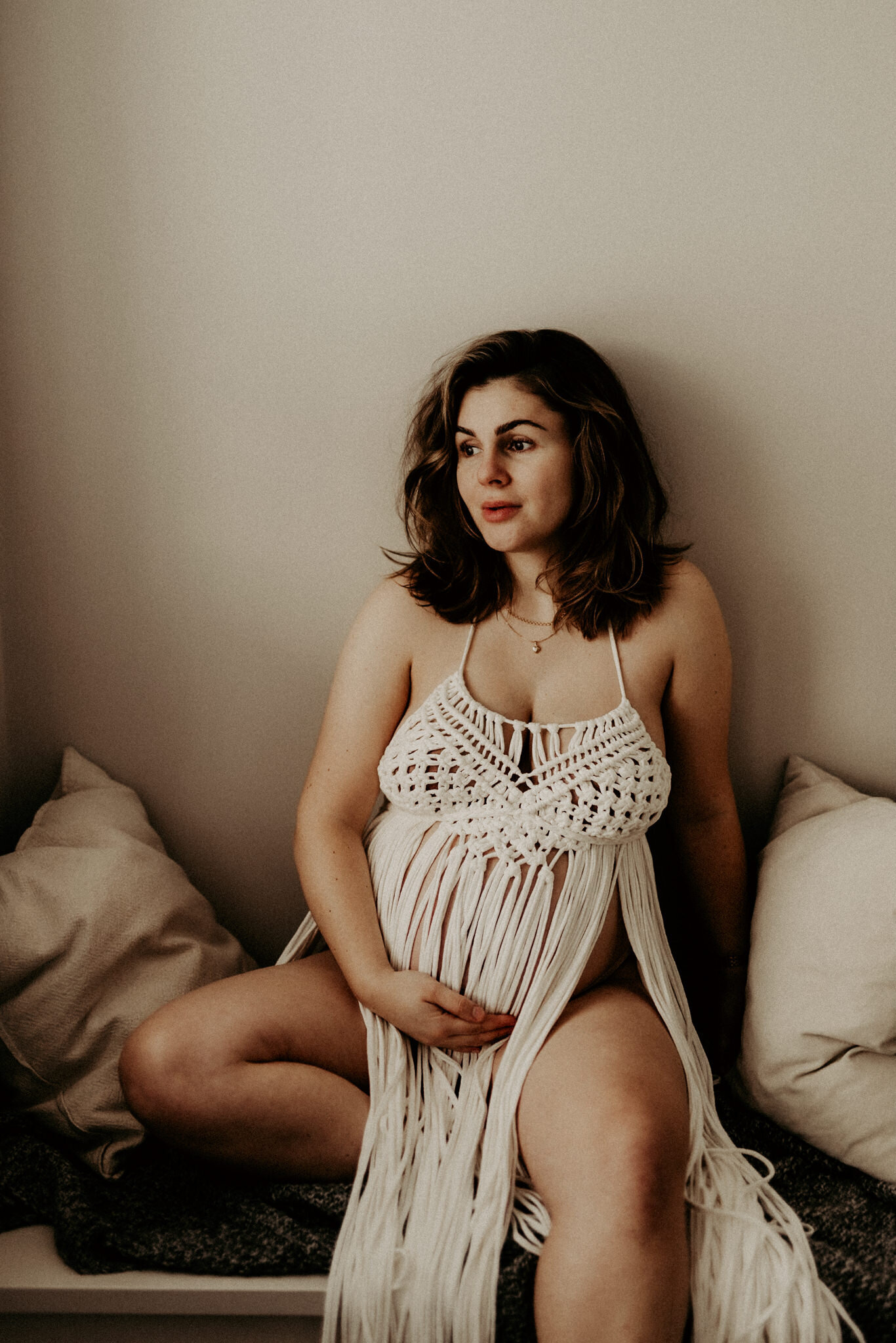 en gravid kvinna sitter stolt och kollar ut genom ett fönster iklädd en gravidklänning i makramé