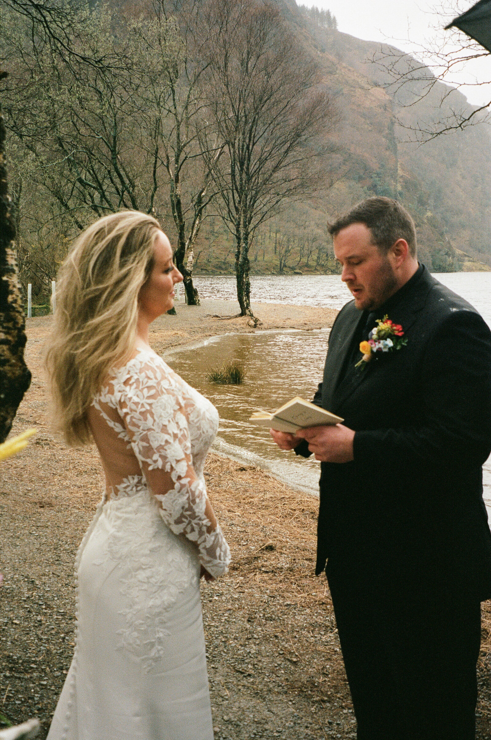 A bride in a lace wedding gown stands beside a man reading from a book in a scenic outdoor setting with a lake and mountains in the background