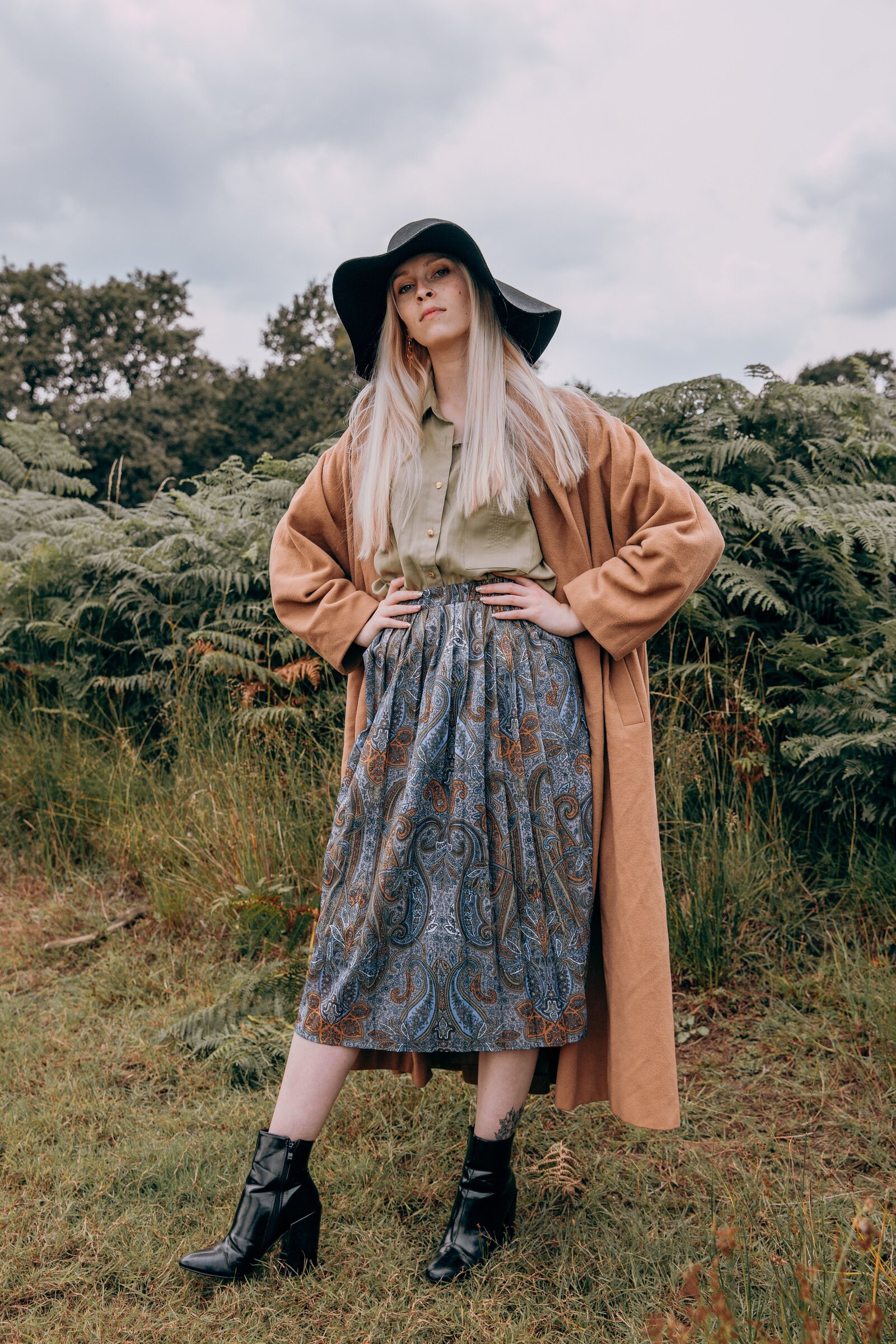 Amber wearing a long brown coat and a blue skirt against a backdrop of ferns in Richmond Park
