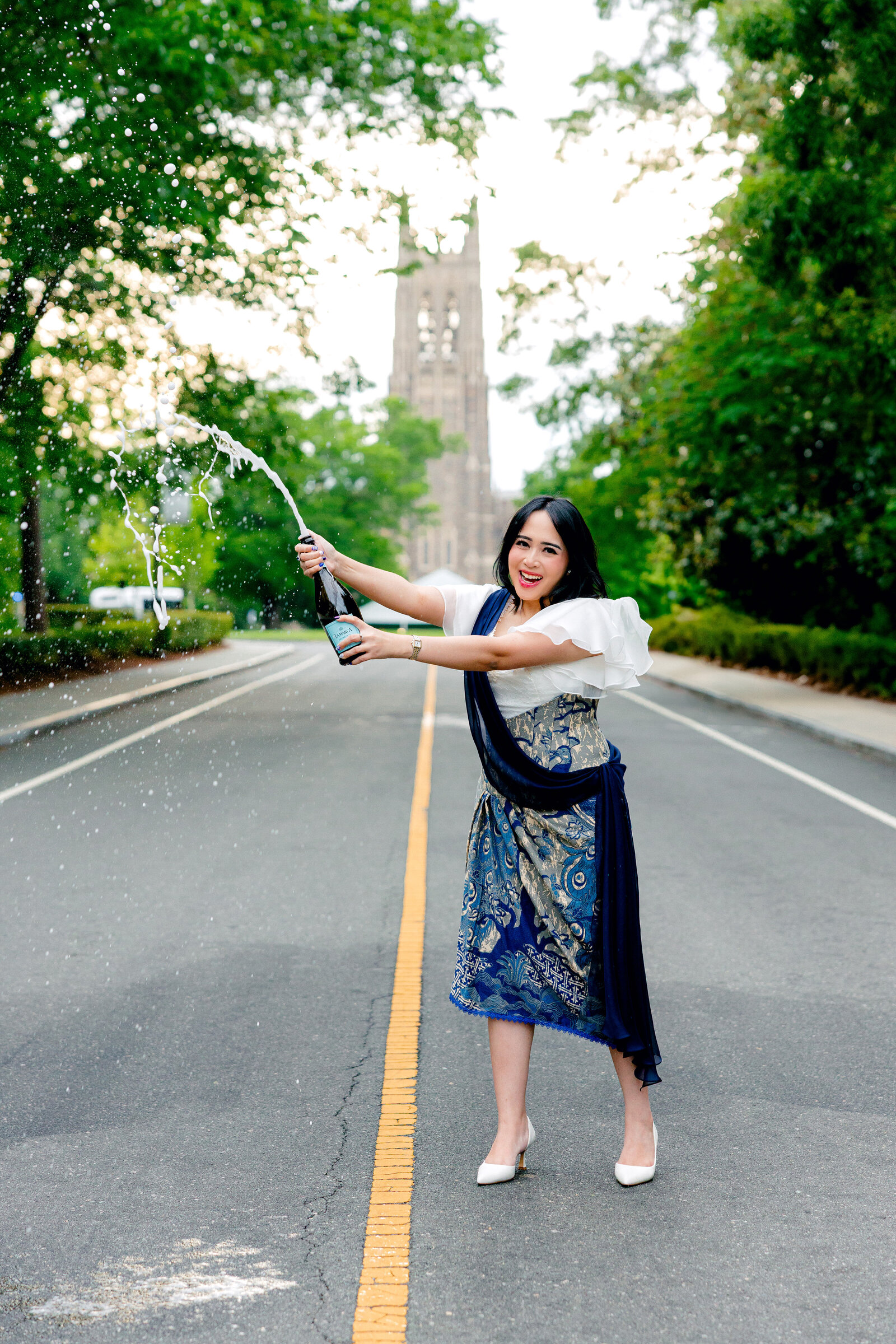 Duke University engineering graduate wearing a traditional kebaya popping champagne on Chapel Drive with Duke Chapel in the background.