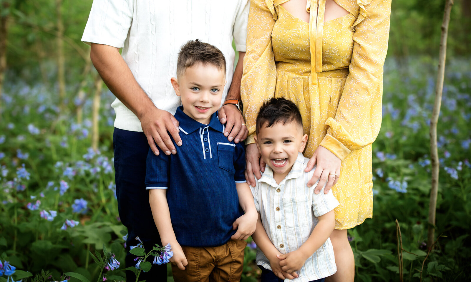Two young boys smile in front of two adults, likely their parents, amid a field of green plants and purple flowers. Capturing the warmth of outdoor portraits, the adults’ faces aren’t visible; one wears a white shirt, the other a yellow dress.