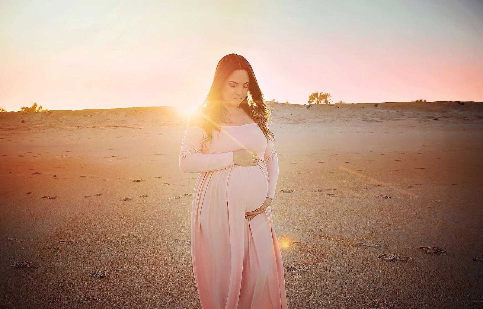A pregnant woman in a pink dress poses on the beach, showcasing the beauty of a maternity session.