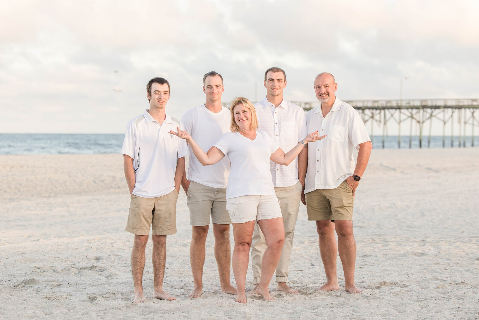 A family of five stands barefoot on the beach near a pier, dressed in coordinated white and khaki outfits. The open sky and gentle ocean waves create a serene backdrop. Ideal for North Carolina family photography, capturing classic beachside portraits.