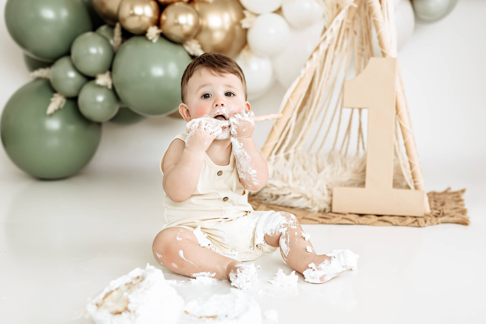 A baby sits on the floor covered in cake and cream, celebrating a first birthday with green and white balloons and a large cardboard number one in the background. The setting is bright and festive.