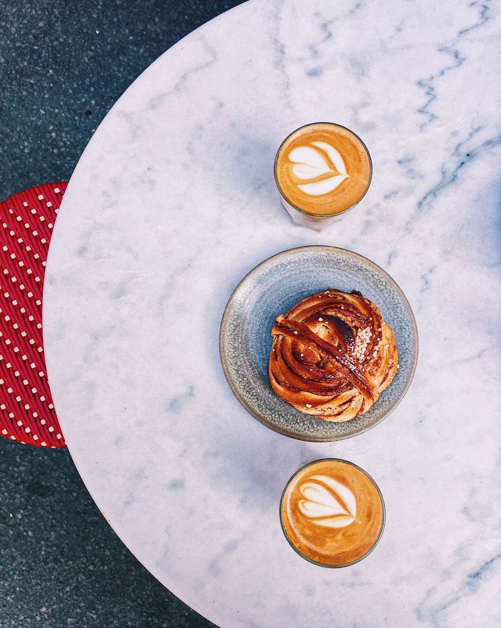 above table view of two cups cappucinos with heart shapes in the foam, and a pastry on a dish in the middle of the two cups.