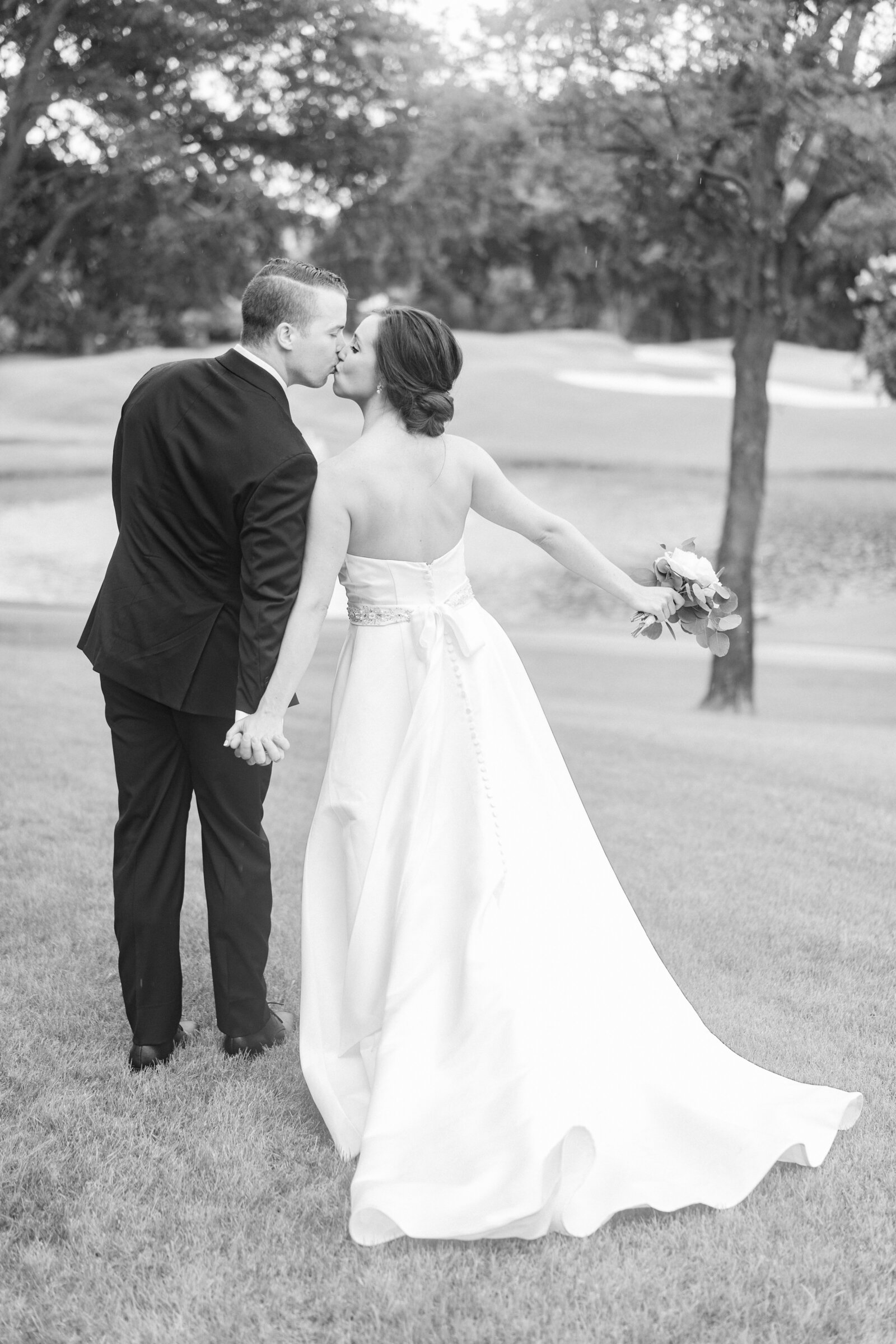 Black and white portrait of a bride and groom in a wedding gown and tuxedo standing on a grass field.