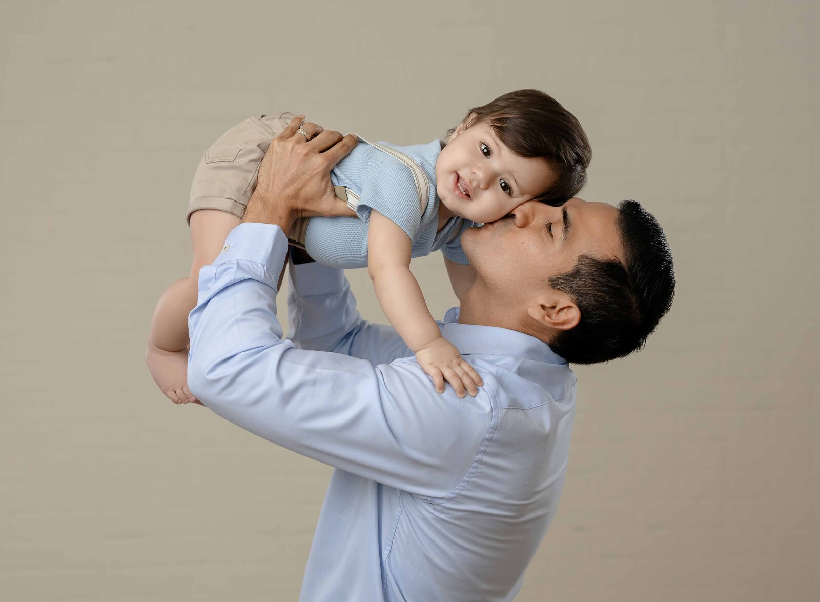 Dad holding baby up in the air and kissing his cheeks. Baby is looking and smiling at the camera.