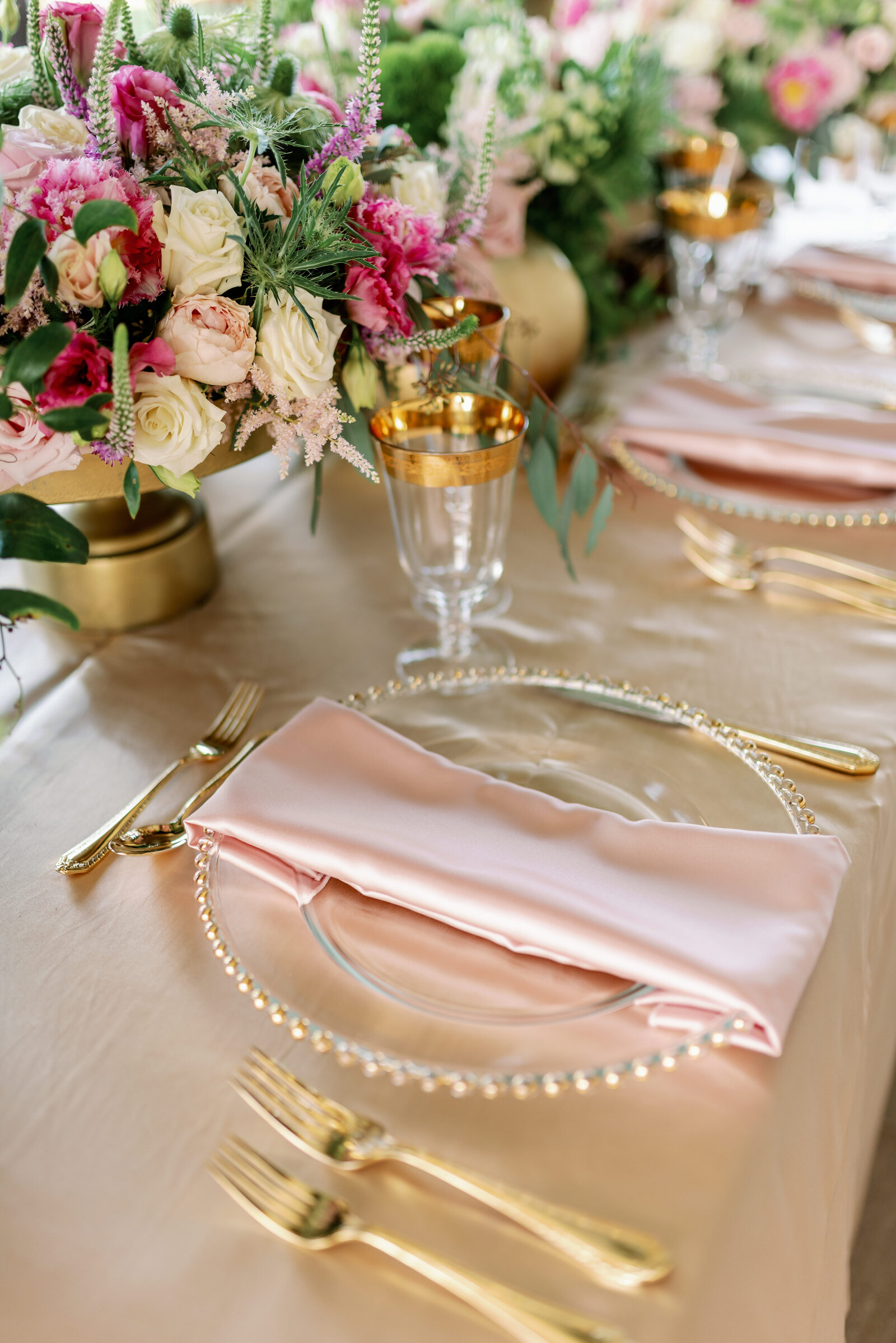 detail of dinner plate with pink silk napkin and gold flatware. A crystal stemmed glass with golden rim sits just above the clear charger plate to the right and a colorful floral arrangement can be seen in the center of the tablescape.