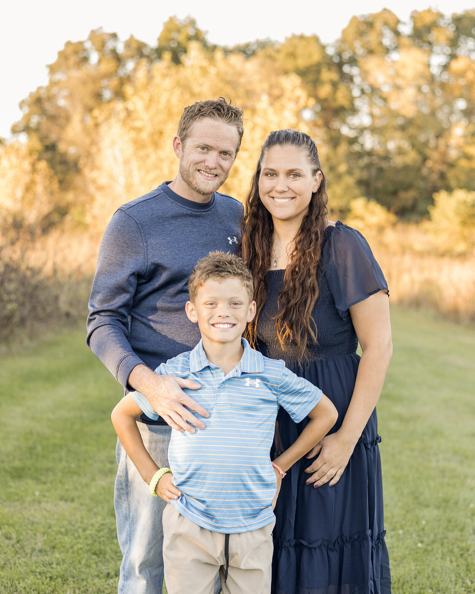 A family of 3 standing in a park.  Dad is wearing a dark blue long sleeve shirt smiling at the camera.  Mom is wearing a dark blue dress smiling at the camera.  Their son is standing in front of them, has sandy blonde hair, smiling at the camera, wearing a light blue and white striped shirt, with his hands on his hips.