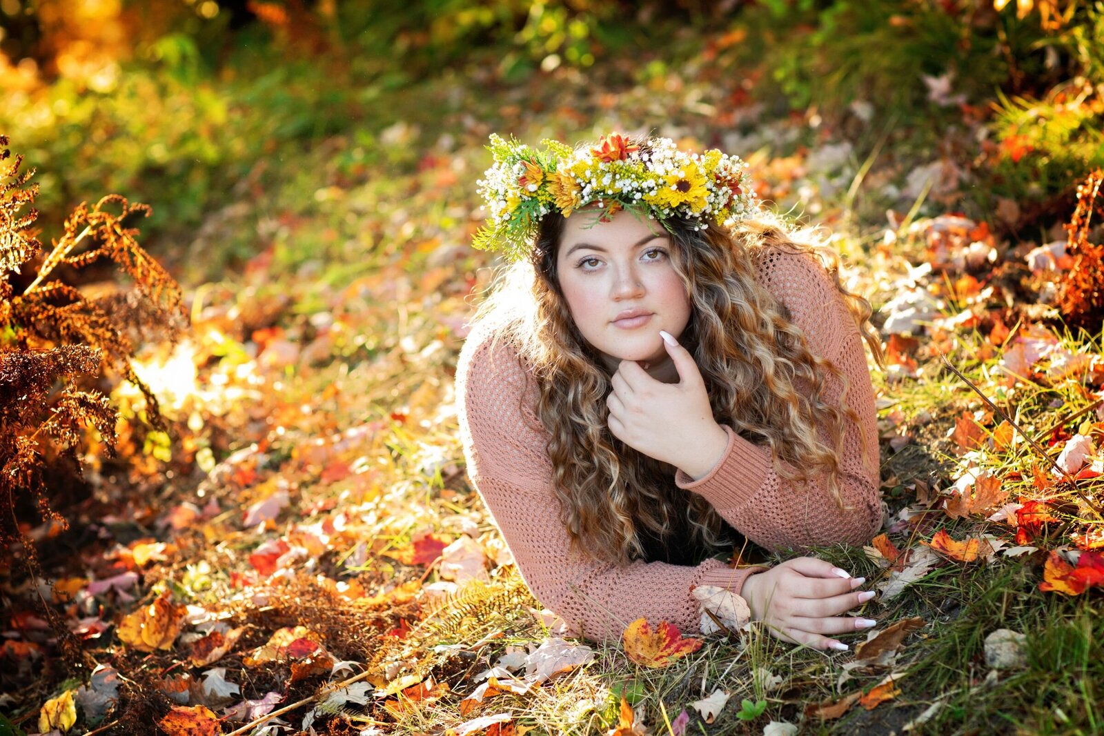 A high school senior girl laying on her stomach  wearing a floral crown with a serious expression with fall colors of amber shine all around her.