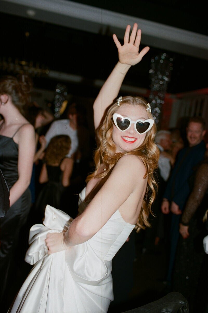 A woman in a strapless white dress with a large bow, wearing heart-shaped sunglasses, smiles and raises her hand at a lively indoor party. She has long wavy hair with pearl clips and red lipstick. People in elegant attire are in the background.
