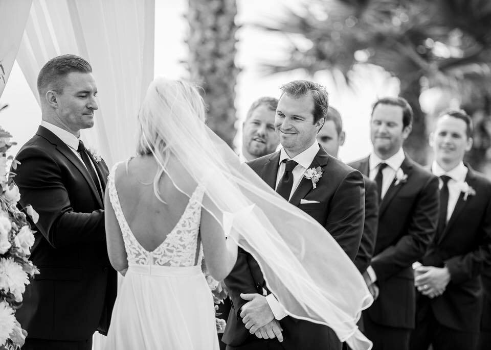 Black and white photo of a wedding ceremony, groom smiling at the bride with the groom's men behind the groom