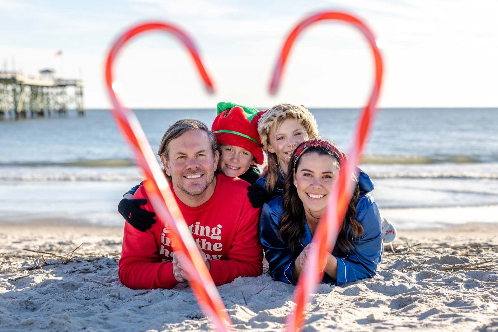 A family of four lies on the sandy beach, framed by a candy cane heart. Dressed in holiday attire, including a Santa hat and festive sweaters, they enjoy a playful moment by the ocean. Perfect for North Carolina family photography, capturing holiday cheer by the sea.