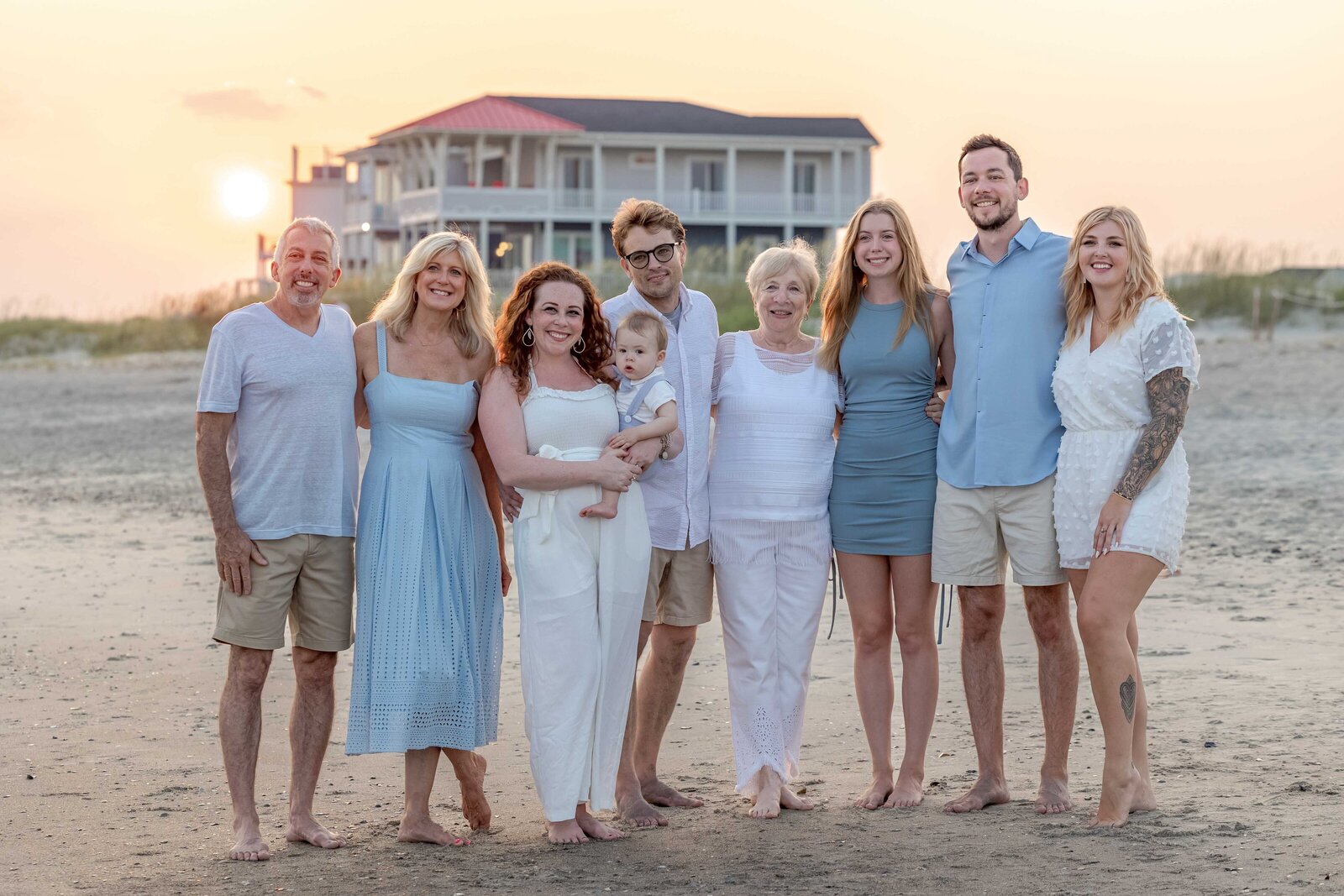  A family poses on the beach at sunset with a beach house in the background. Dressed in pastel and white, they enjoy the serene coastal scenery. Perfect for North Carolina family photography, capturing warm and elegant beachside moments.