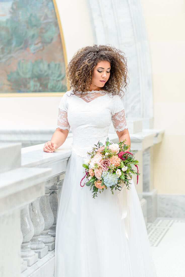 A bride in a white lace wedding dress holds a colorful bouquet on the second floor of the Utah State Capitol Building. Captured by Salt Lake Photographer Melissa Woodruff Photography