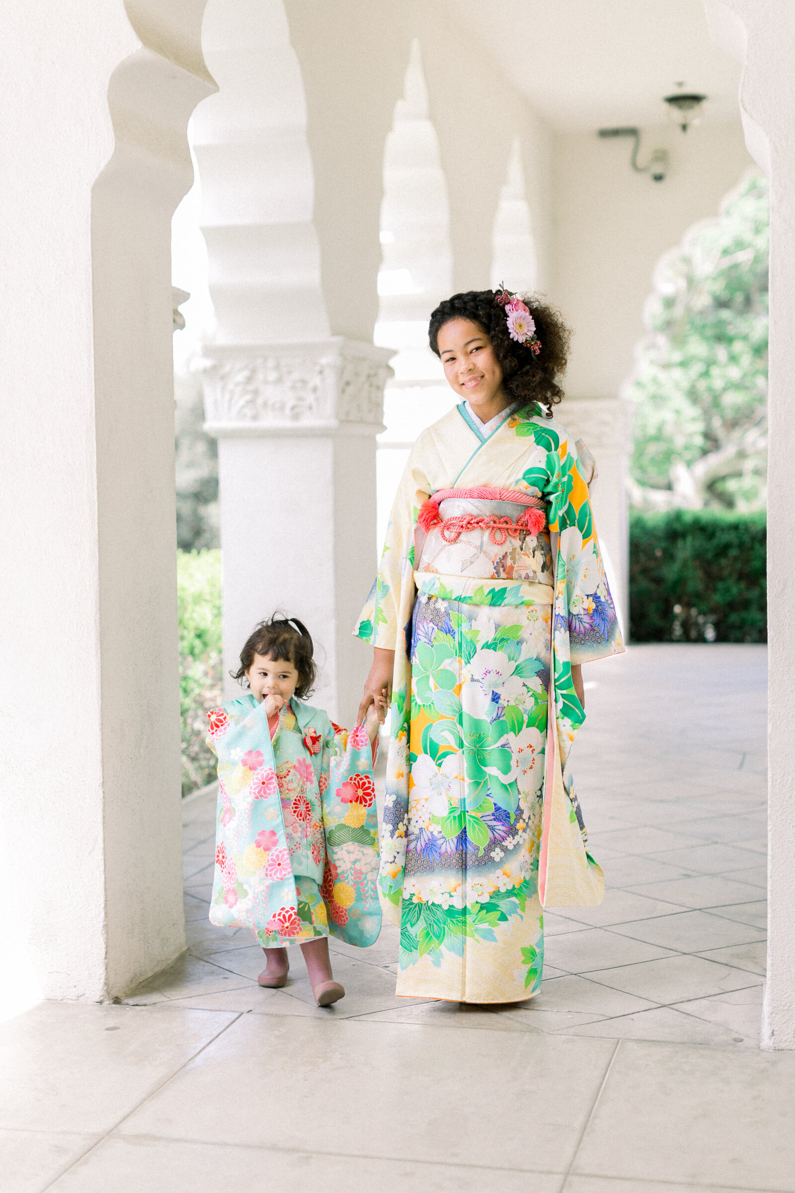 Older sister in a floral furisode kimono walking hand in hand with a little sister in matching kimono during Seijinshiki at Brand Park Glendale, kimono rental Los Angeles | ロサンゼルス・ブランドパークでの成人式、振袖姿の姉と可愛い着物の妹が手をつないで歩く姿
