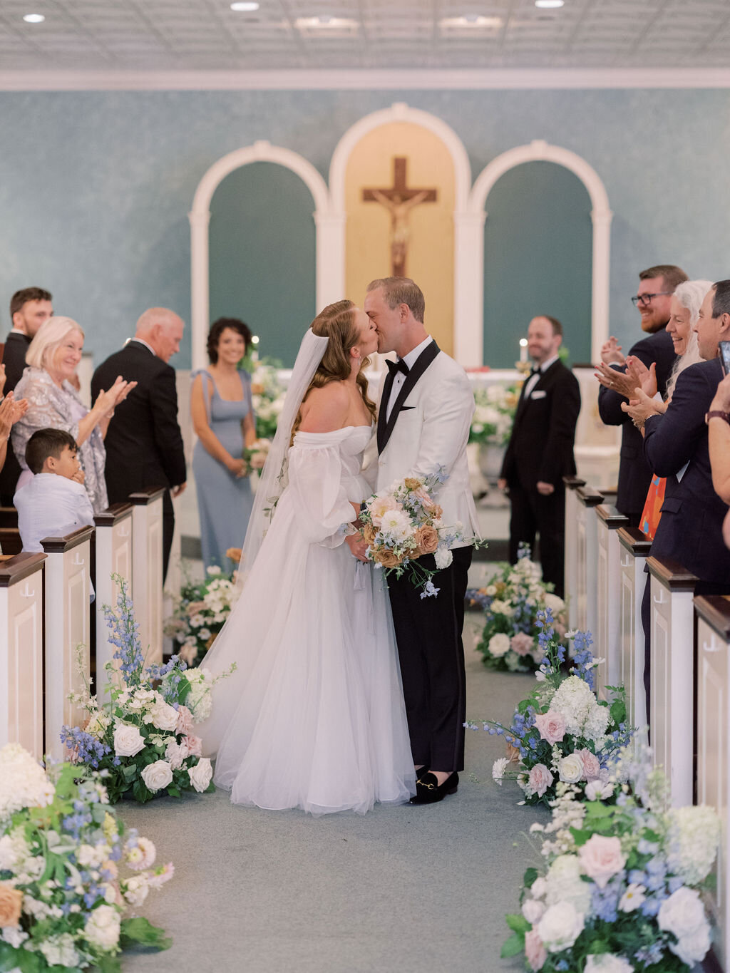Bride and groom kissing in the Sacred Heart Catholic Church in Chestertown MD aisle, framed by a meadow of floral arrangements down the aisle including blue delphinium, white hydrangea, blush roses, white dahlias and toffee roses.