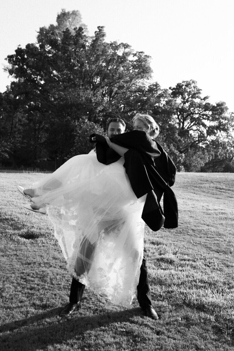 A groom lifts the bride playfully in a grassy field. They are both smiling, and the brides dress flows in the breeze. Trees stand in the background on a sunny day. The photo is in black and white.