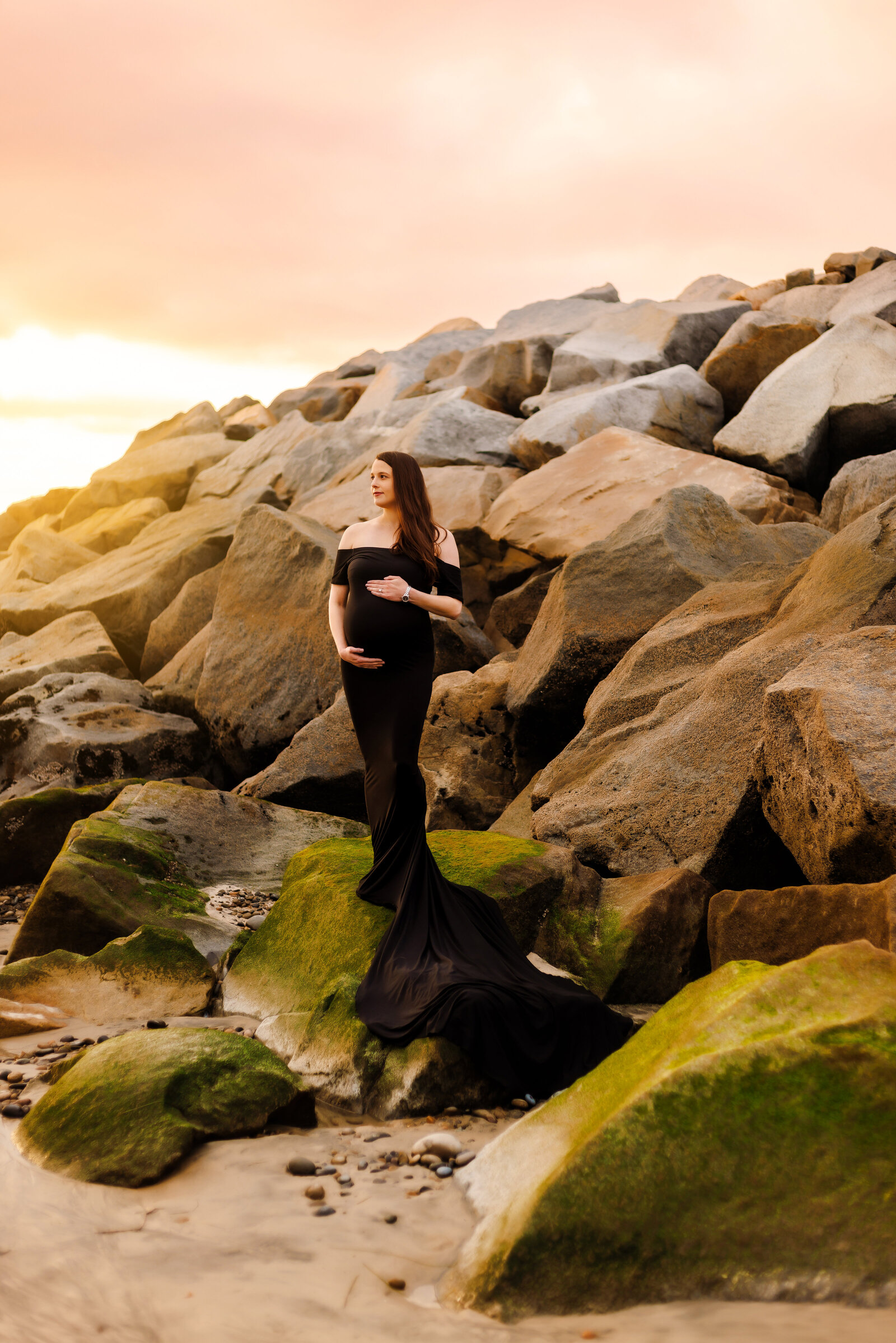 Maternity Photographer, an expectant mother stands  on beach rocks in a gown
